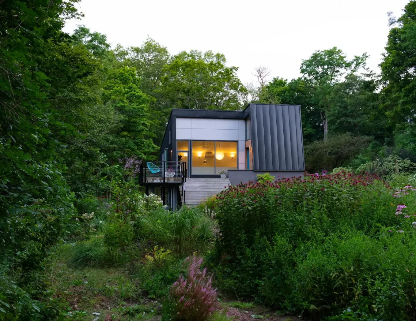 Modern dark-clad container structure with illuminated windows nestled in lush woodland garden setting by noroof architects.