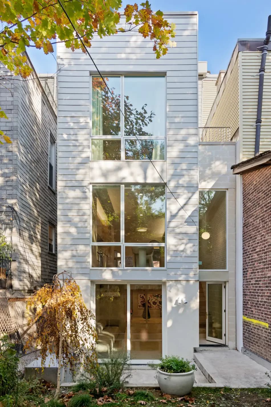 Tall narrow townhouse facade with white shiplap siding and full-height gridded windows, flanked by brick buildings, by noroof architects.