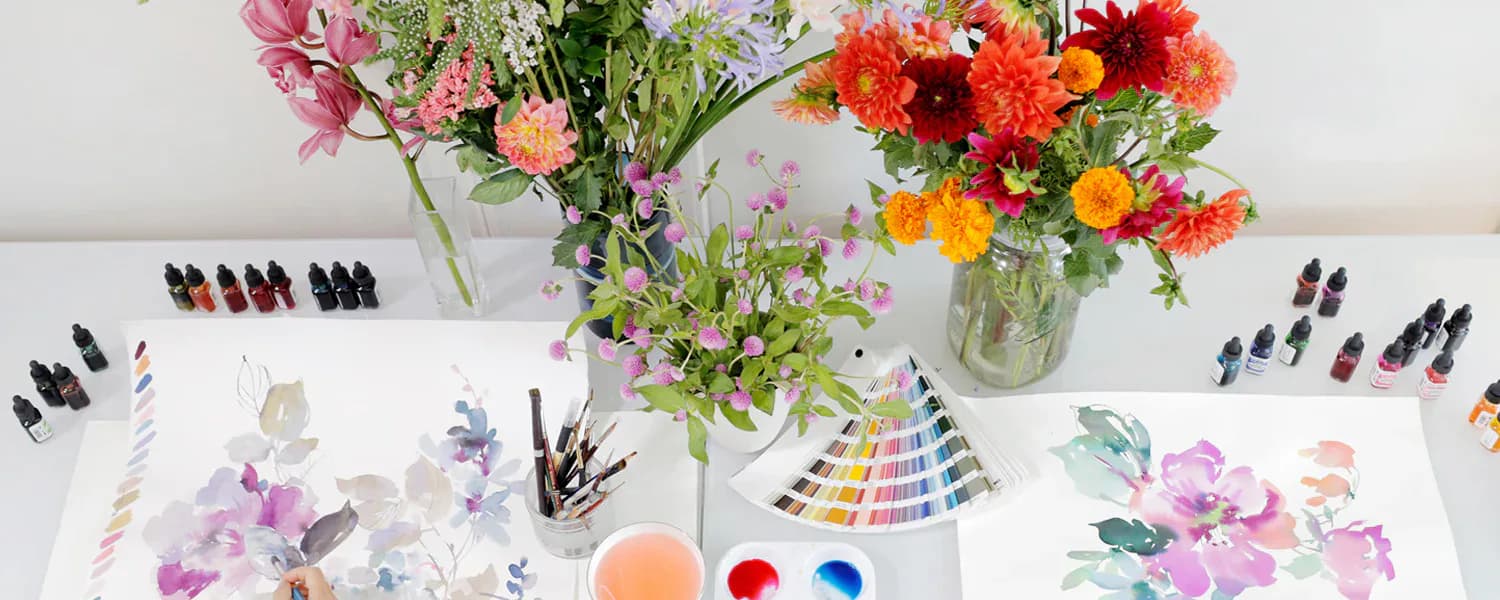 Flat lay of watercolor palette, paint swatches, and fresh floral arrangements in glass vases displaying botanical design work by NuPrimary.