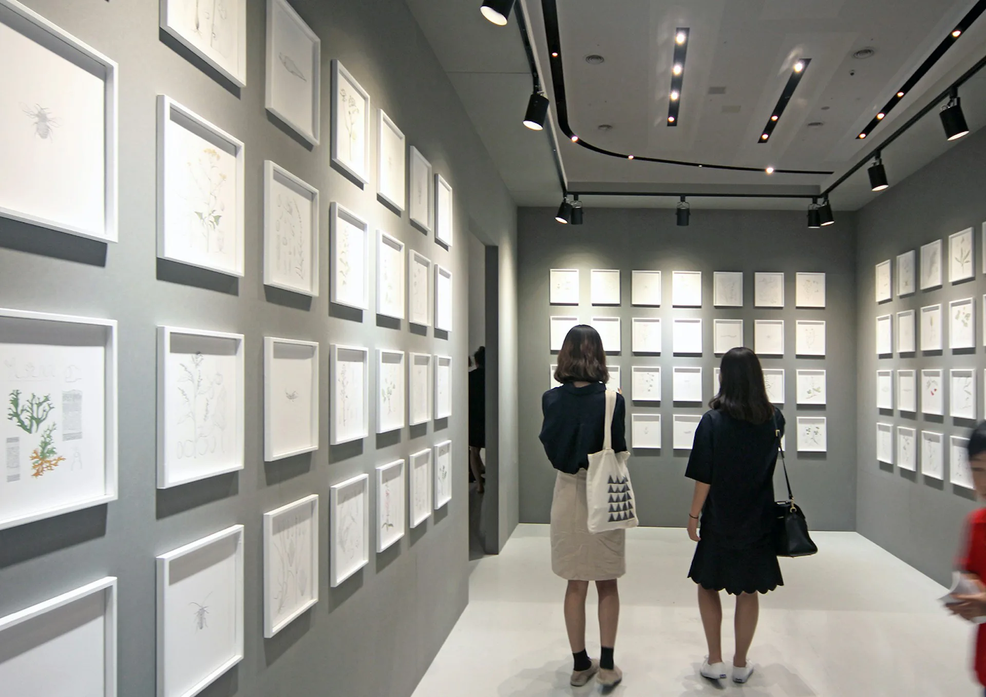 Gallery interior with two visitors viewing illuminated white framed artworks on gray walls under track lighting, minimal aesthetic by OBRA Architects.