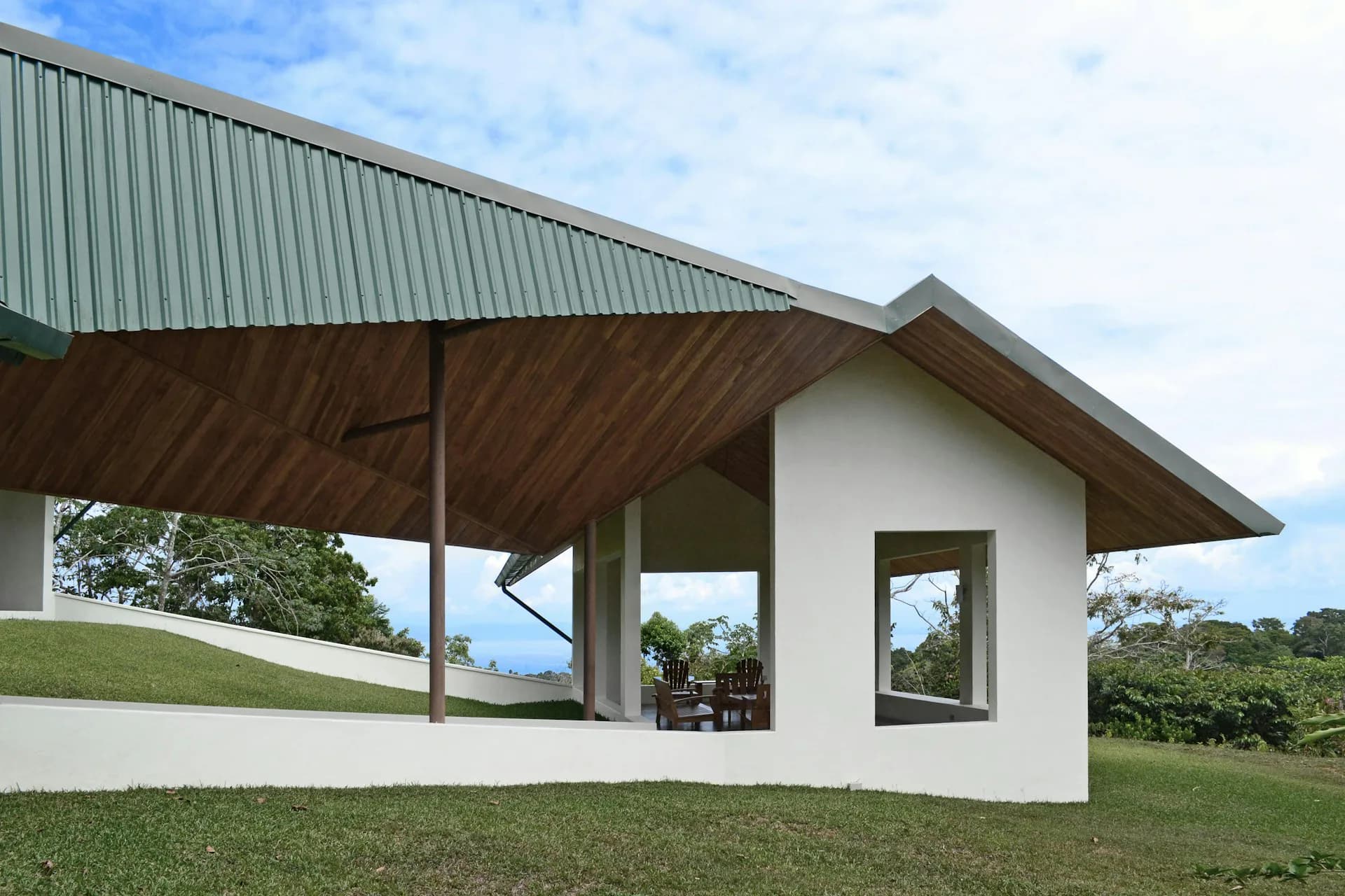 Modern residential pavilion with white walls, wood ceiling, metal roof, and open-air covered terrace overlooking green landscape by OBRA Architects.