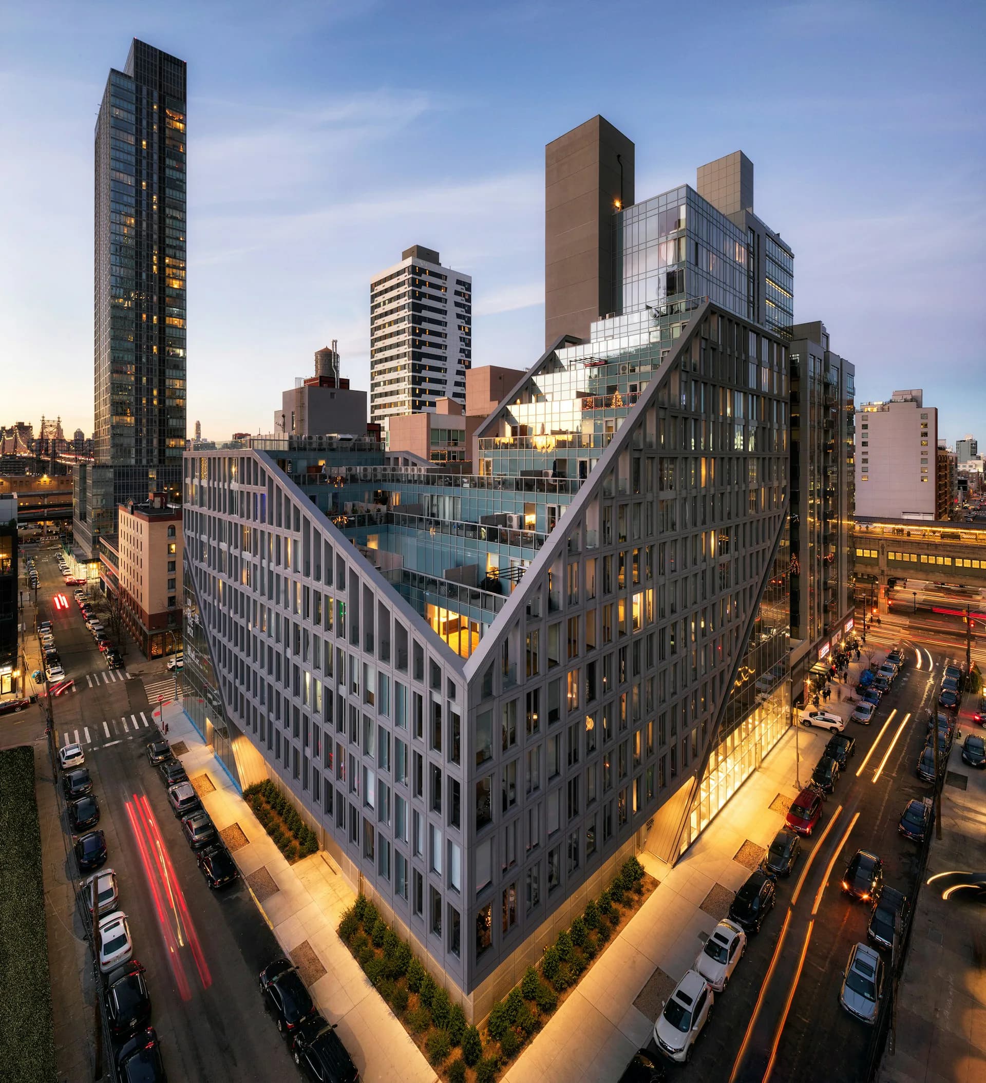 Corner view of a modern mixed-use building with glass and brick facade, illuminated at dusk, surrounded by Manhattan streetscape and neighboring buildings by ODA.