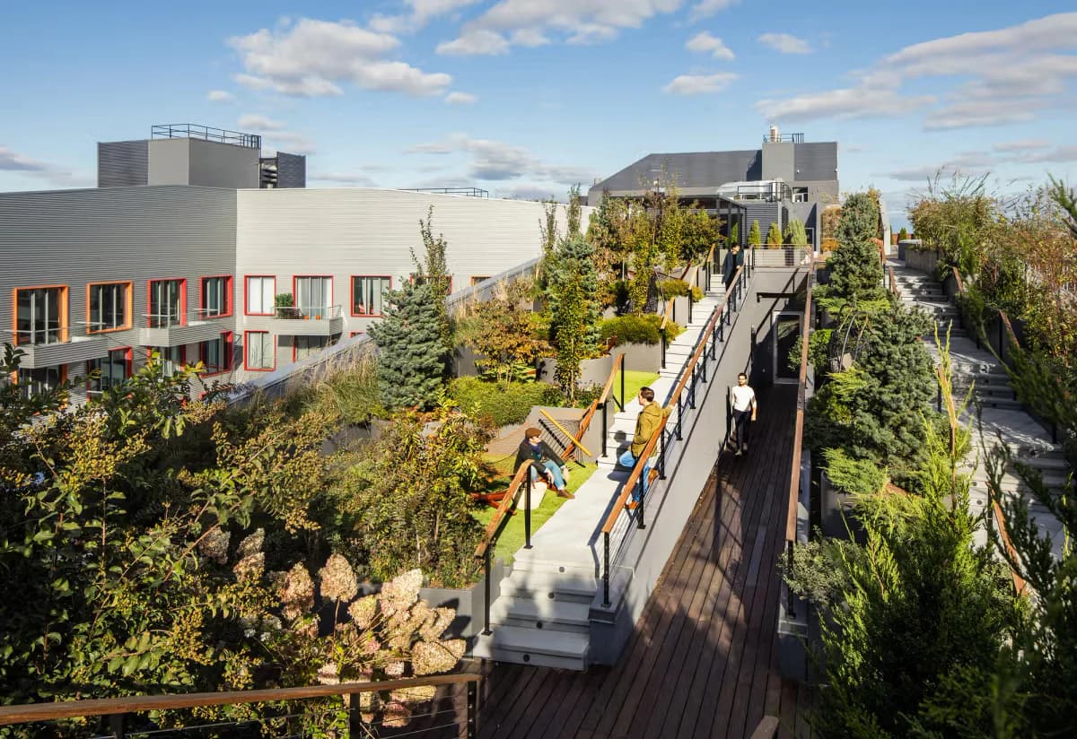 Elevated wooden pedestrian bridge with railings overlooking landscaped grounds and modern building complex with orange accents by ODA New York.