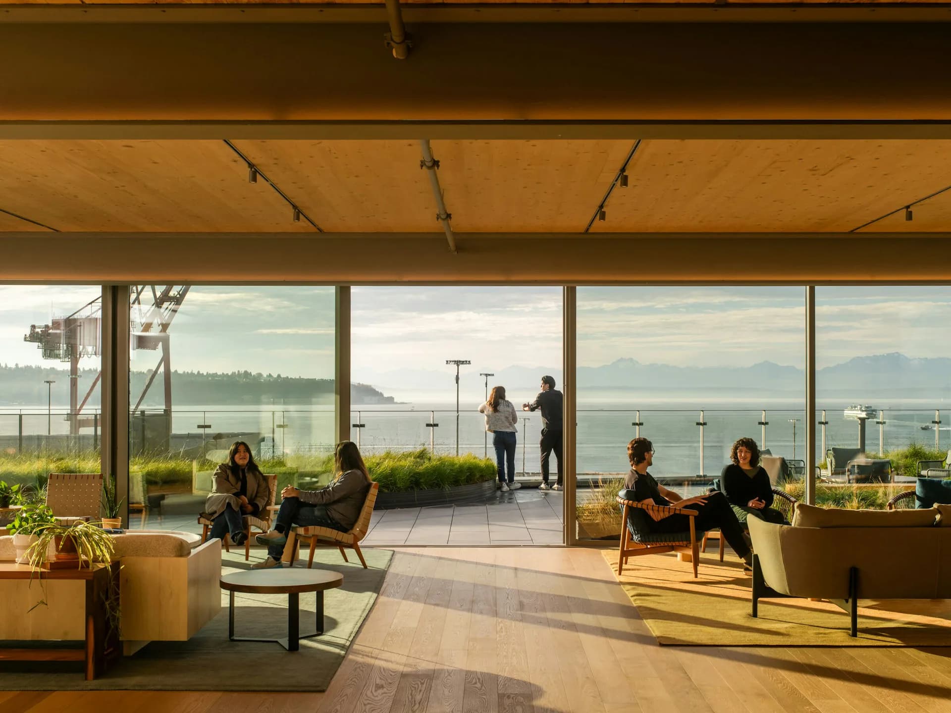 Modern office lounge with floor-to-ceiling windows overlooking mountains, featuring mid-century chairs, warm wood ceiling, and natural light by Olson Kundig.
