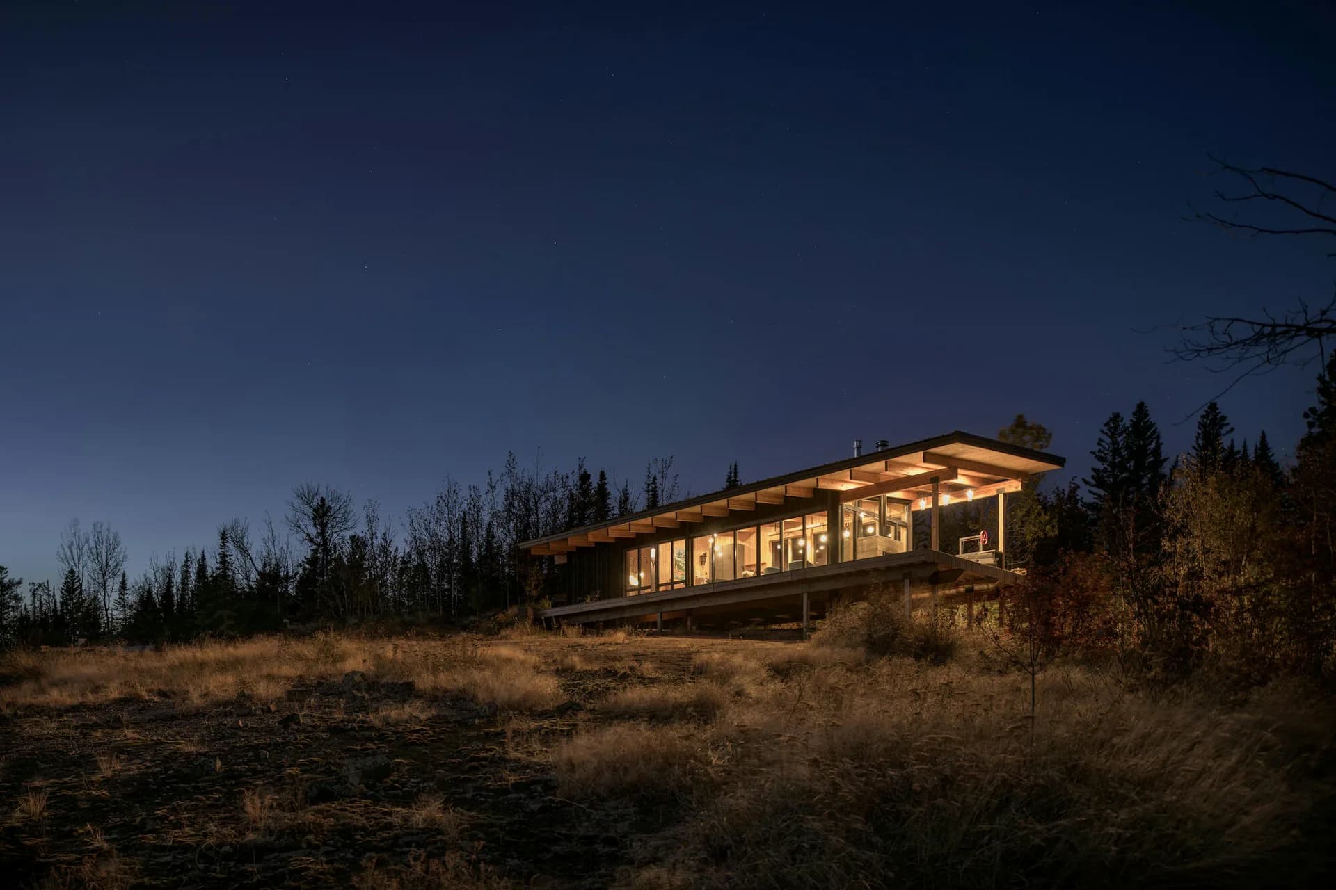 Modern residential structure with expansive glazing illuminated at dusk, nestled in a forested landscape with dry vegetation by Olson Kundig.