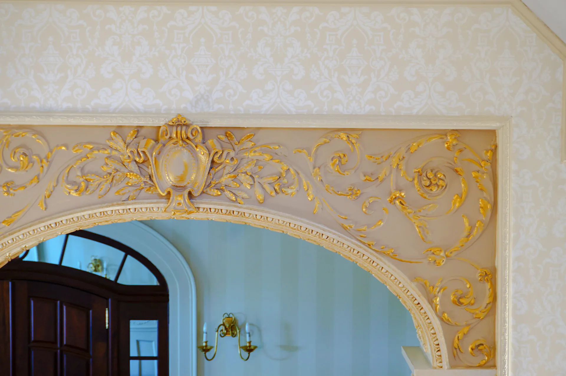 Ornate archway with gilded baroque detailing and cream-colored wallpaper framing a doorway with classical wall sconces by Open Shop Arts.