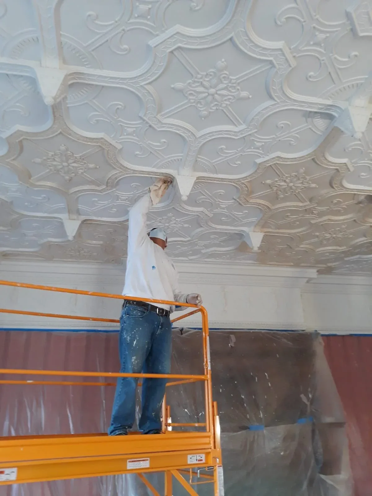 Worker on scissor lift installing ornate decorative plasterwork ceiling with intricate molded medallions and floral details by Open Shop Arts.