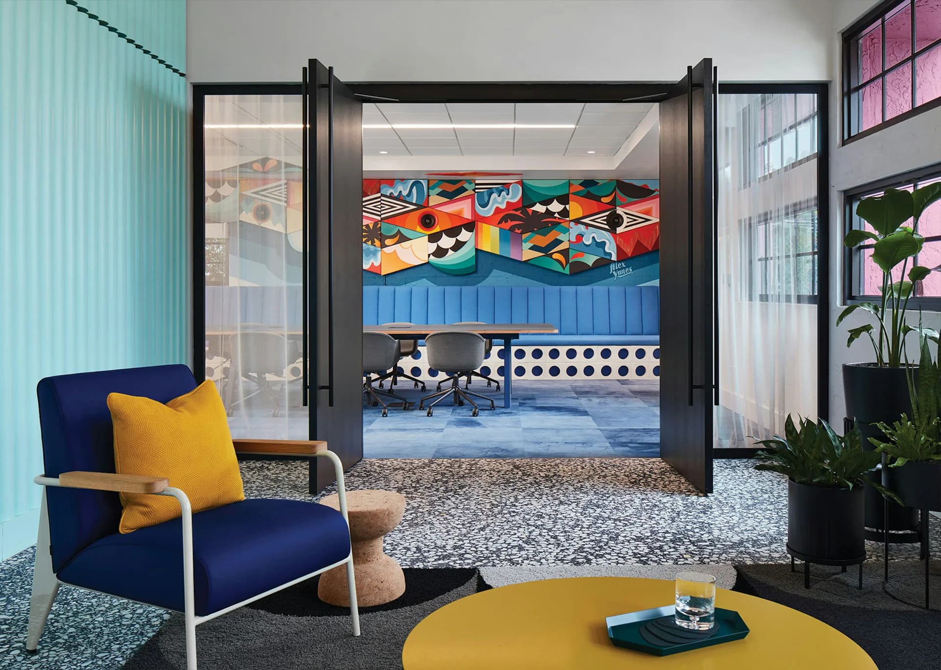 Modern office lounge with navy armchair, yellow accents, and colorful play area visible through glass doors by OTJ Architects.