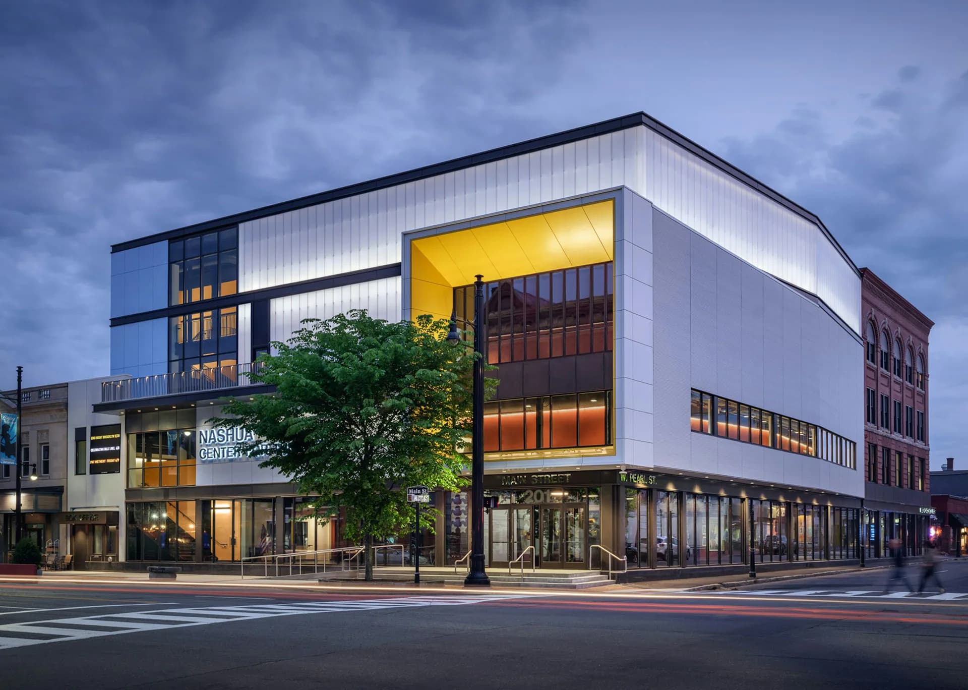 Contemporary commercial building with white metal paneling, yellow-lit upper level, and glass storefront at dusk, designed by OTJ Architects.