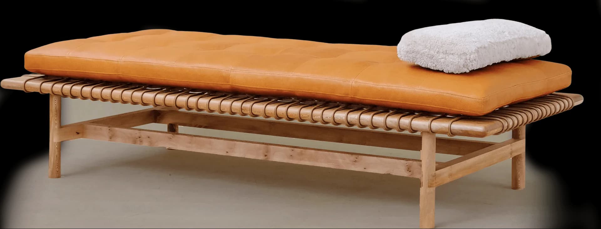 Minimalist wooden daybed with woven rope detailing, tan leather cushion, and gray pillow on natural wood frame by Otzi.