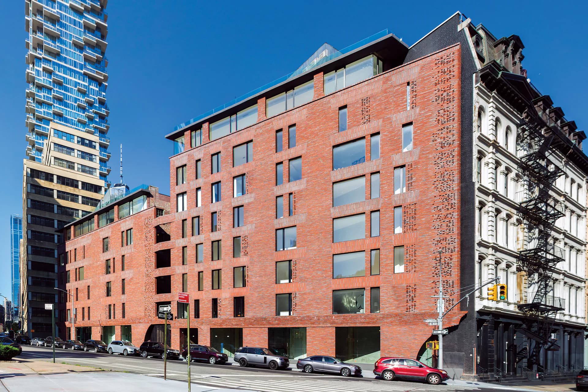 Red brick mid-rise residential building with large windows and ground-floor retail, situated among modern towers under clear blue sky, by Palette Architecture.