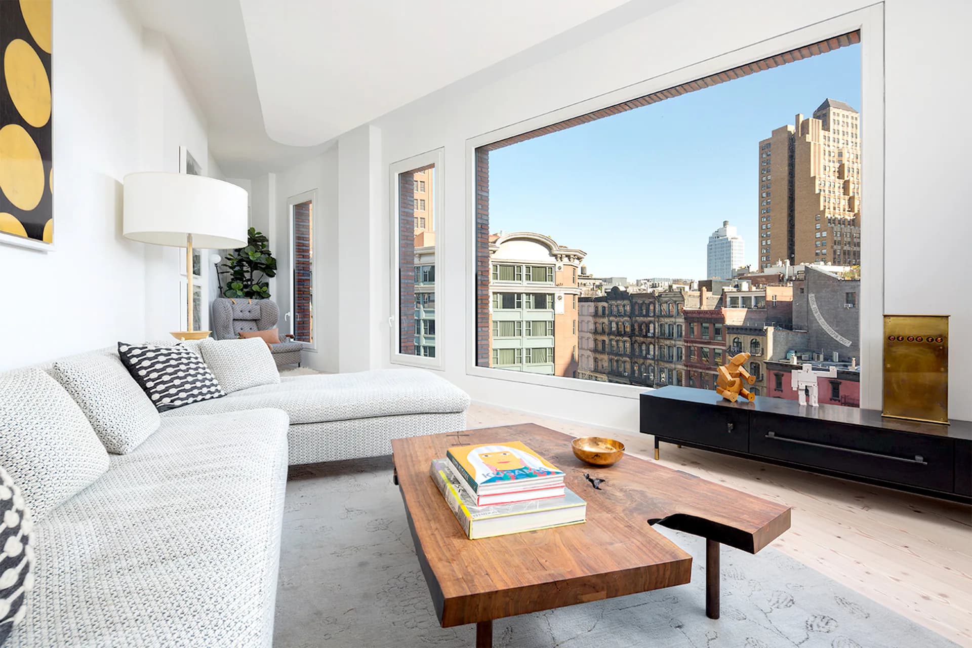 Bright modern living room with floor-to-ceiling windows overlooking Manhattan skyline, white furnishings, wood coffee table, and geometric artwork by Palette Architecture.