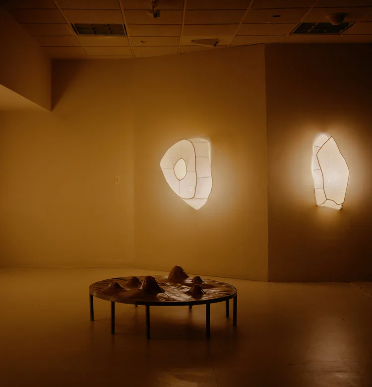 Warm-toned gallery installation with abstract geometric light fixtures on walls and a sculptural table with organic forms by Panorammma.