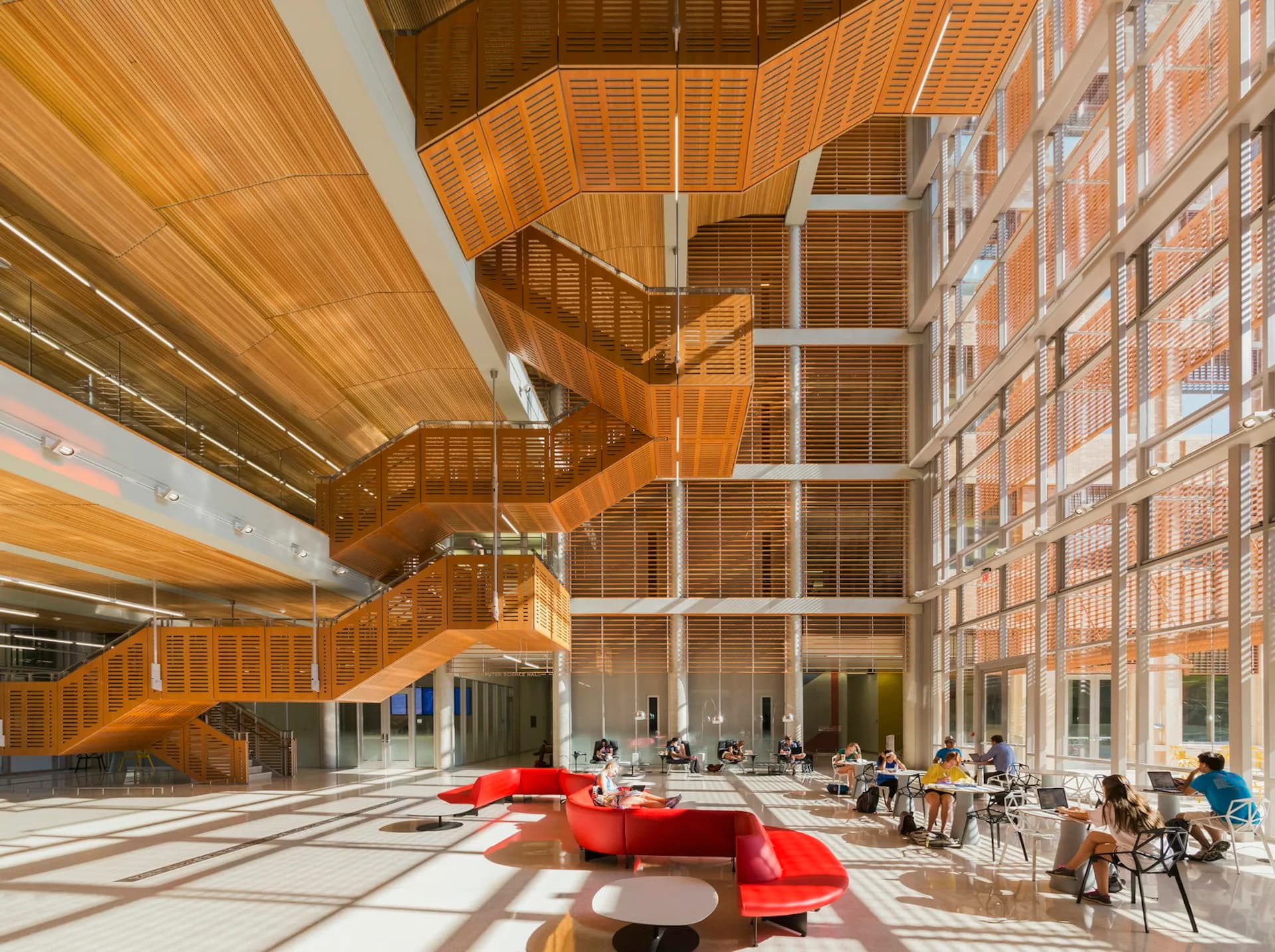 A modern atrium with dramatic wooden ceiling elements, brick facade, and red seating areas bathed in natural light, designed by Pelli Clarke & Partners.