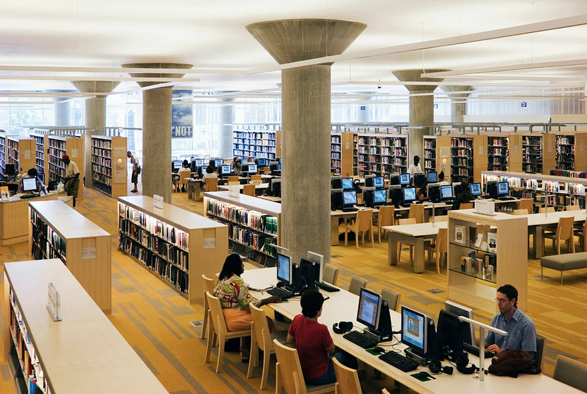Modern library interior with wooden floors, exposed concrete columns, natural light, and computer workstations integrated among bookshelves, designed by Pelli Clarke & Partners.
