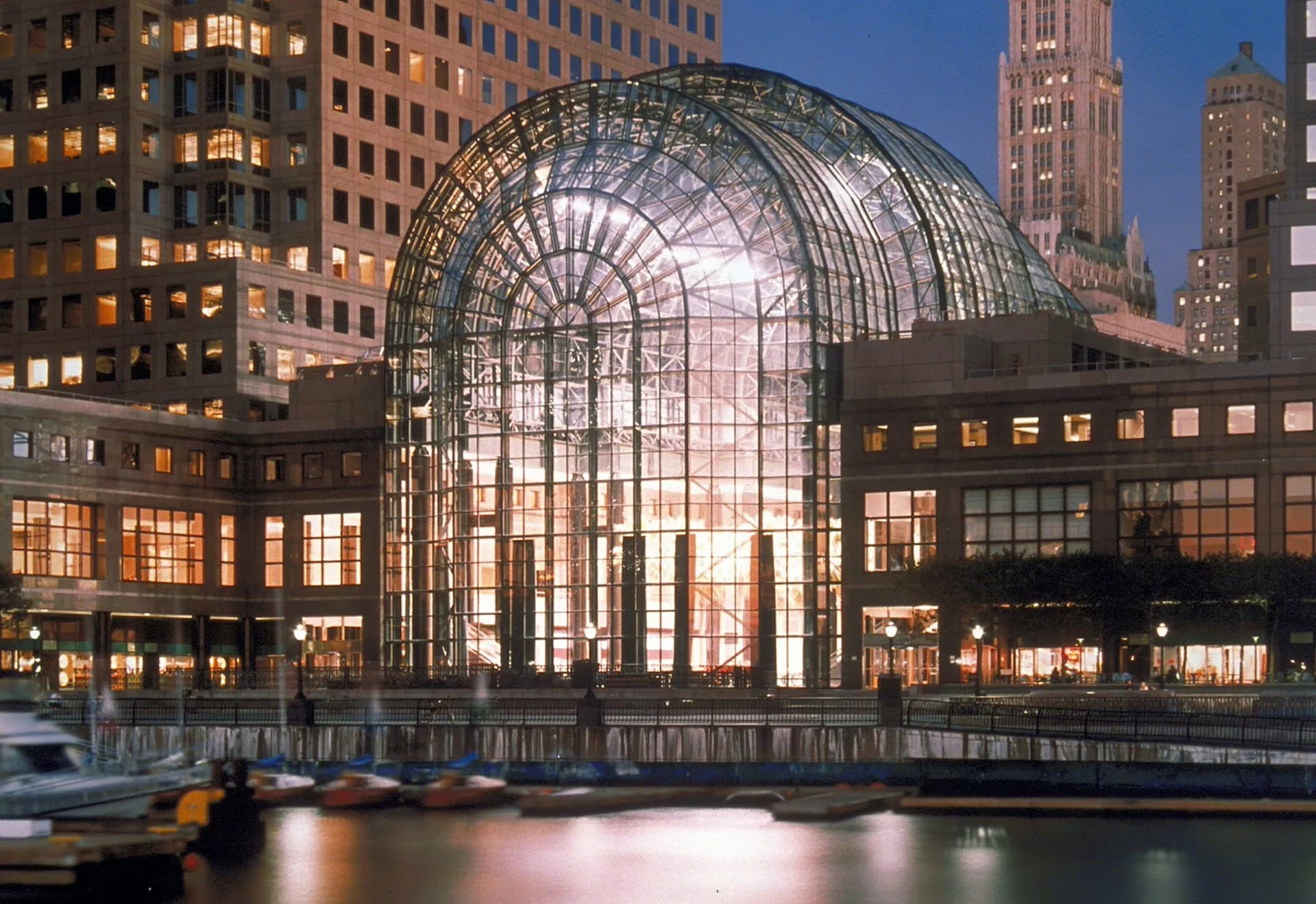 Illuminated glass and steel arched transit terminal at dusk with surrounding Manhattan skyscrapers by Pelli Clarke & Partners.