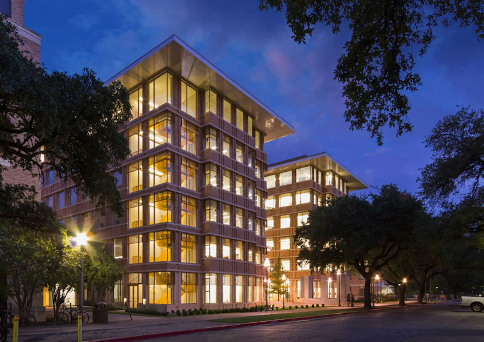 Illuminated institutional buildings with golden-lit façades and large gridded windows at dusk, surrounded by mature trees, by Pelli Clarke & Partners.