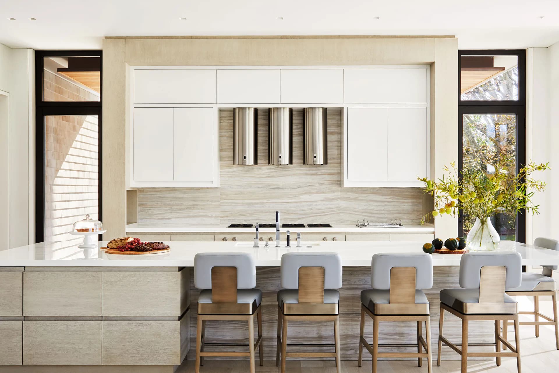 Bright modern kitchen with white cabinetry, light wood backsplash, waterfall island with seating, and natural light from large windows by Pembrooke & Ives.