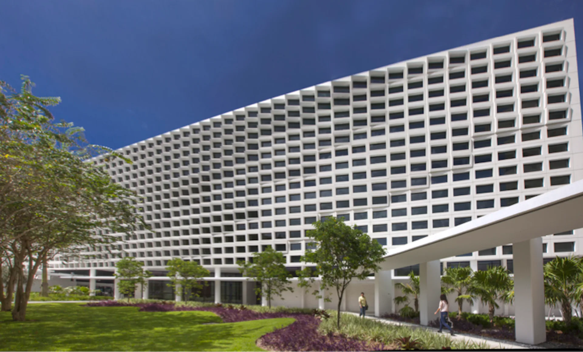 Modern curved multi-story hotel or residential building with repetitive white grid facade, ground-level landscaping with trees and lawn, and covered colonnade by Perkins&Will.