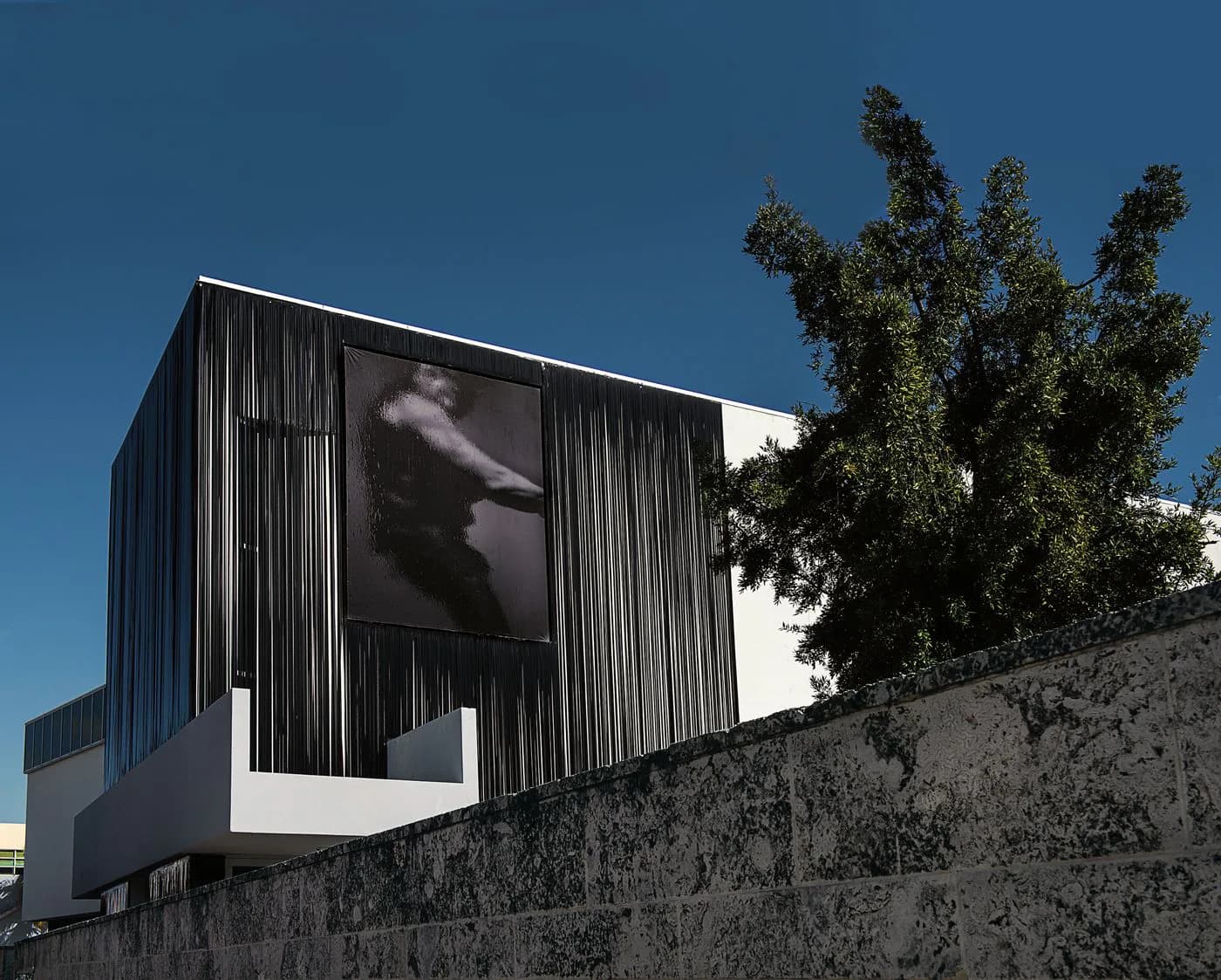 Modern black corrugated metal building with large window display overlooking concrete wall and green tree, architectural design by Peter Marino Architect.