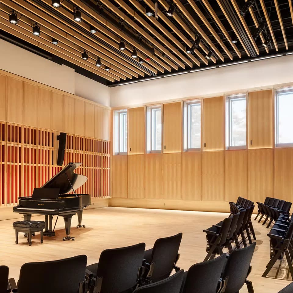 Modern music recital hall with wooden wall paneling, grand piano, tiered seating, and architectural ceiling with integrated lighting by PHT Lighting Design.