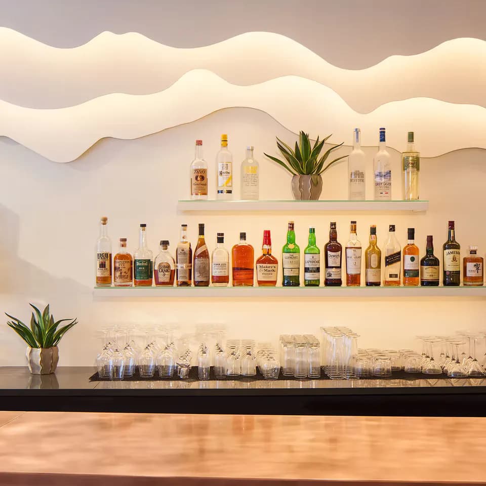 Minimalist bar interior with floating shelves displaying spirits and glassware against a sculptural wave-textured wall, lit by PHT Lighting Design.