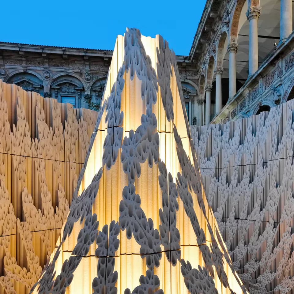 Dramatic architectural installation featuring a tall yellow and dark striped structure in a courtyard with classical buildings, designed lighting creating striking shadow patterns by PHT Lighting Design.
