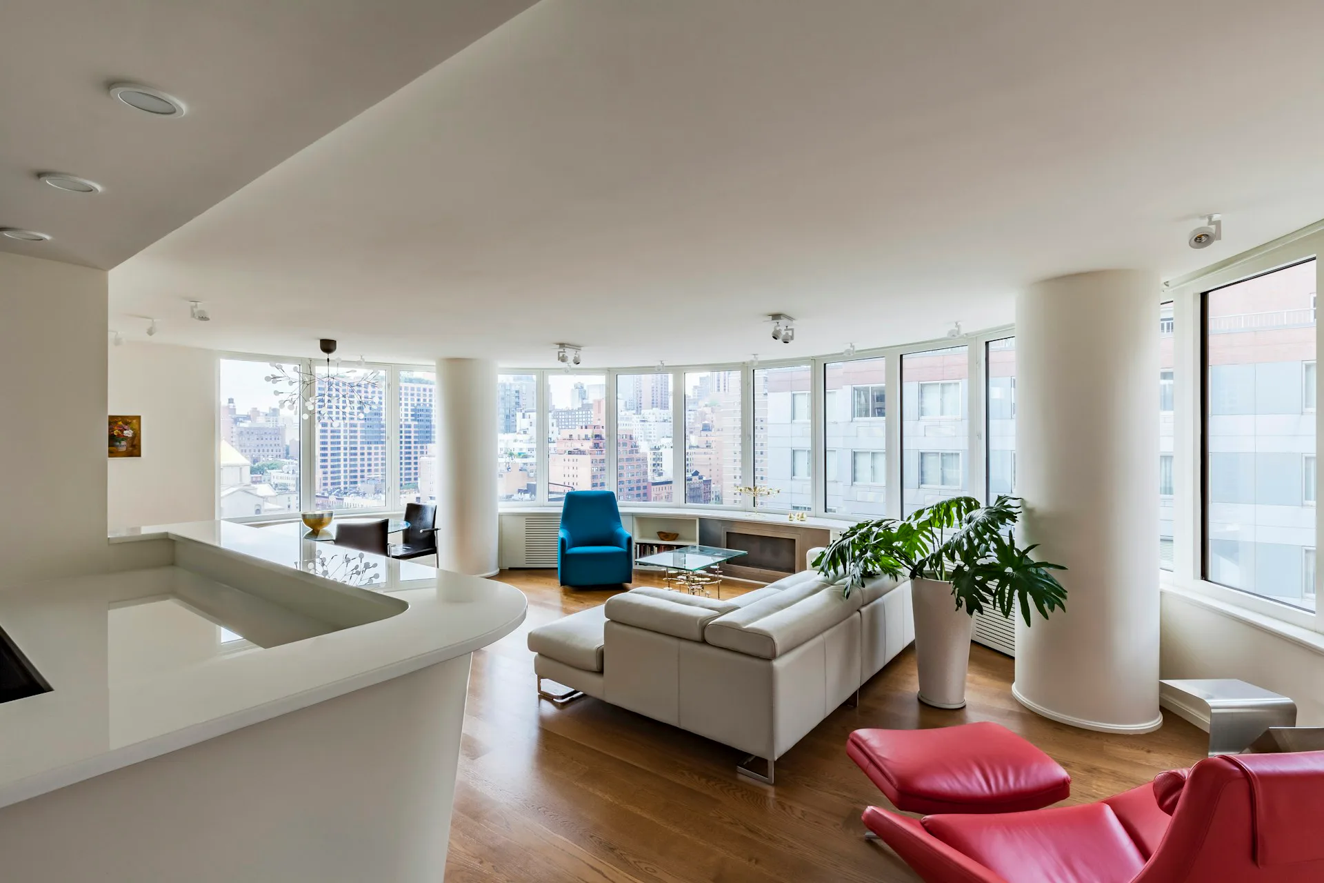 Modern high-rise apartment living room with floor-to-ceiling windows, white kitchen island, blue accent chair, red seating, wooden floors, and city views, designed by Pier, Fine Associates.