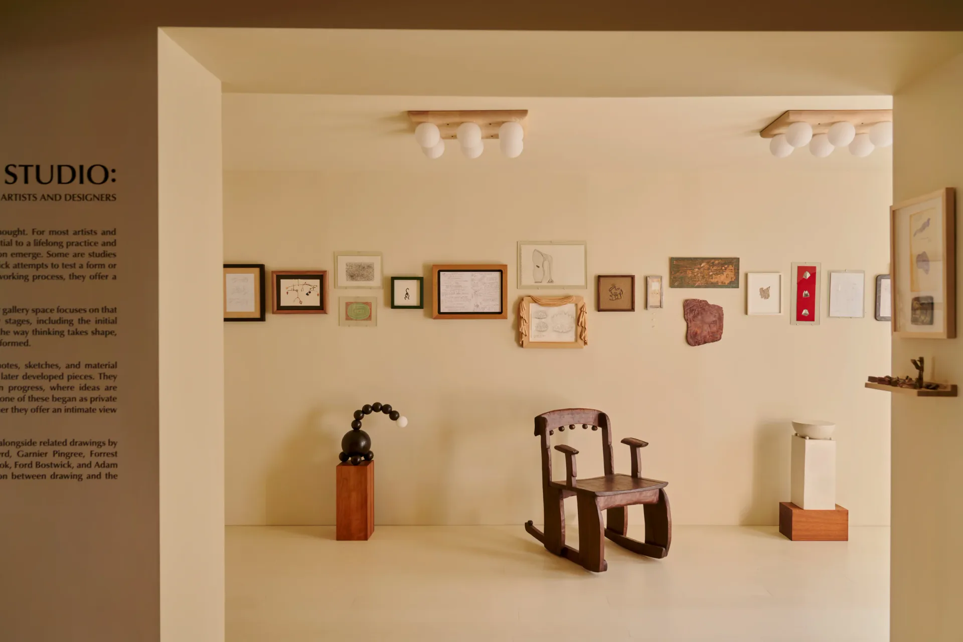 Minimalist studio installation featuring a cream-colored wall with framed artworks, wooden furniture pieces, and sculptural objects arranged on a white floor by Piscina.