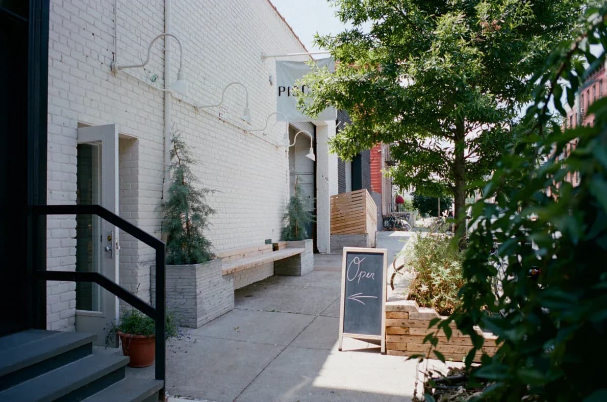Sunlit courtyard with white brick walls, wooden doors, climbing ivy, and potted plants, creating an intimate European-style outdoor space by Piscina.
