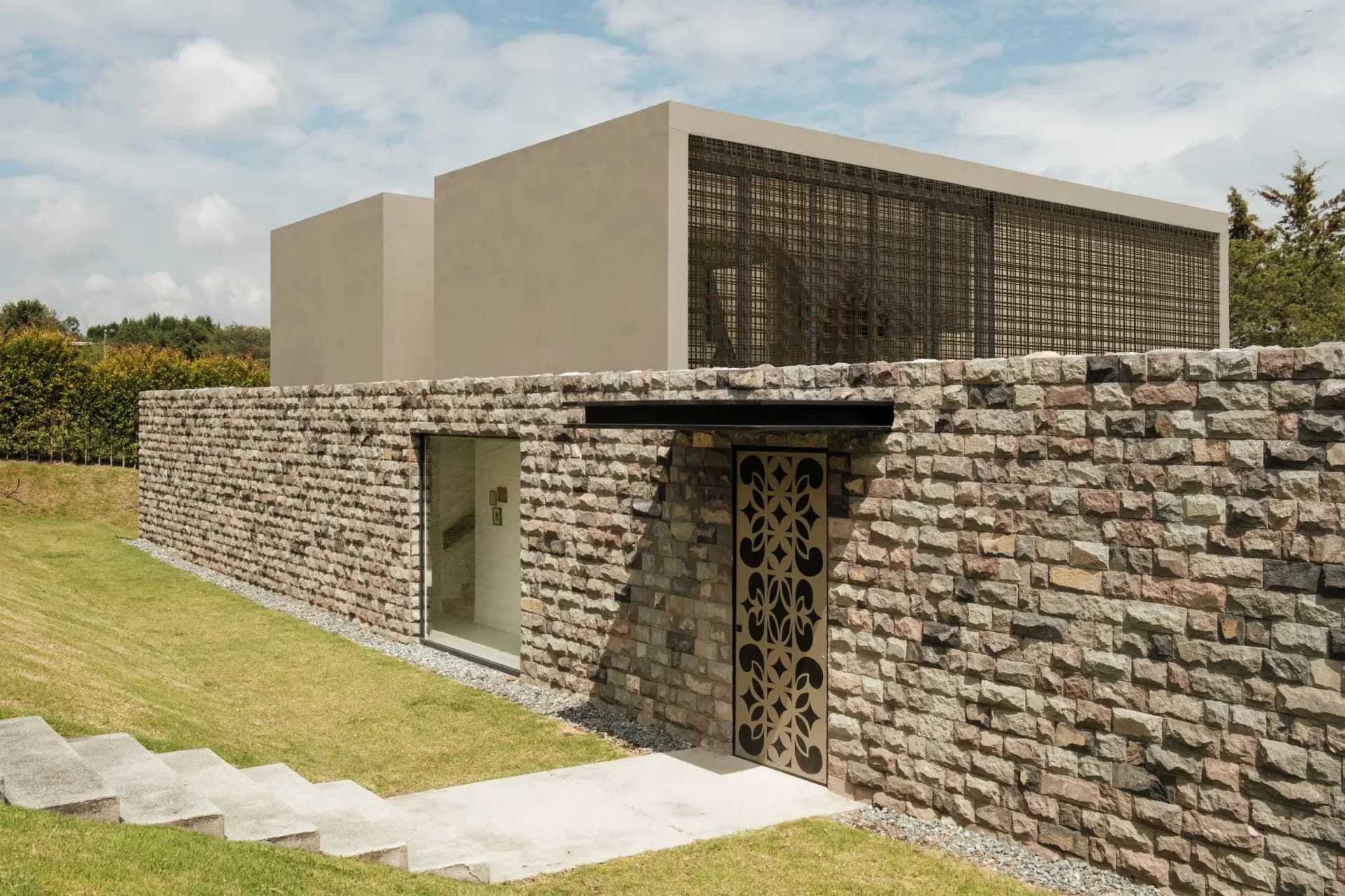 Contemporary residential building with stone base, concrete upper volume, and metal screening, situated on grassy landscape by PJCArchitecture.