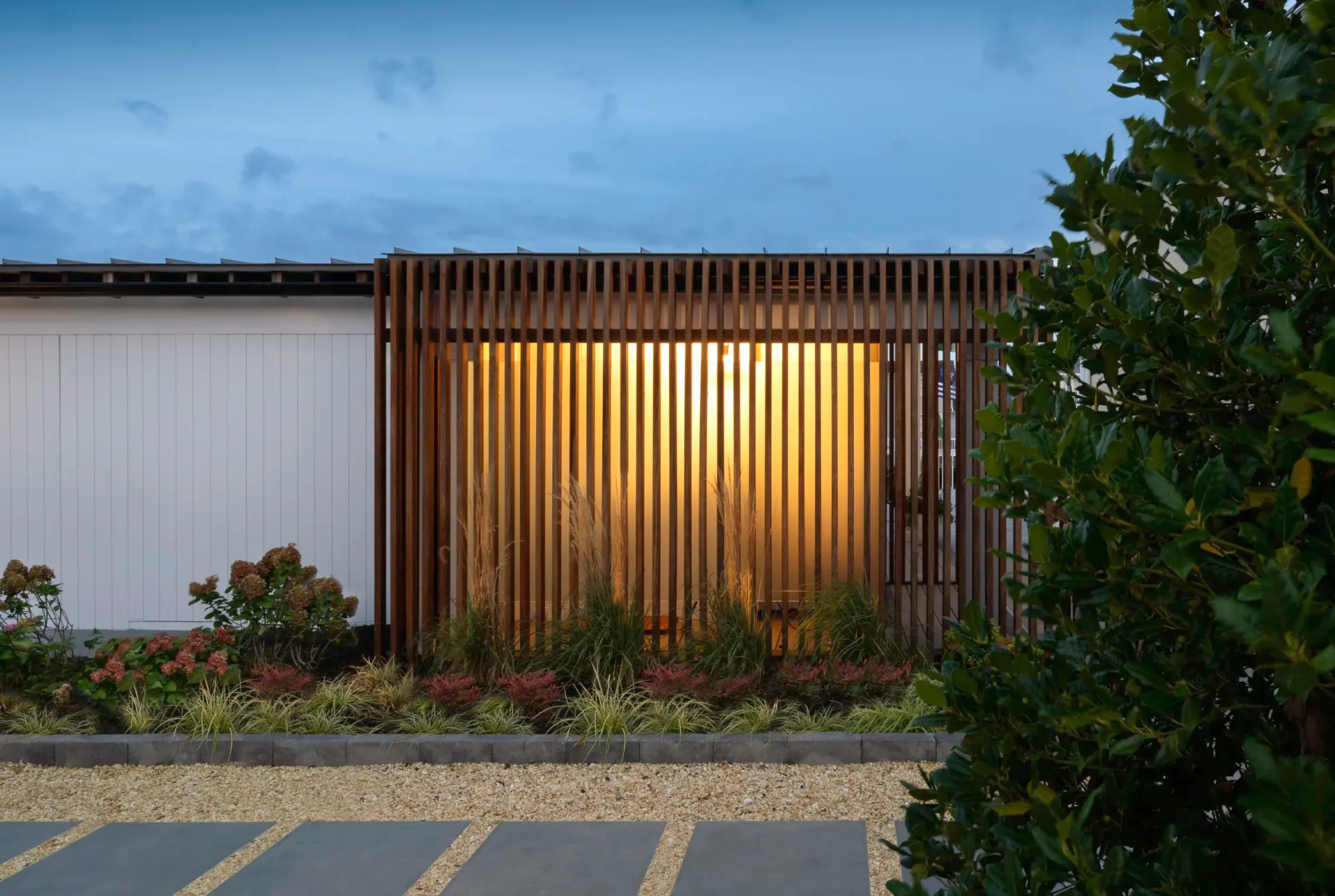 Modern residential facade with vertical wood slat screening and warm interior lighting, minimal landscaping, and adjacent white wall structure by PJCArchitecture.