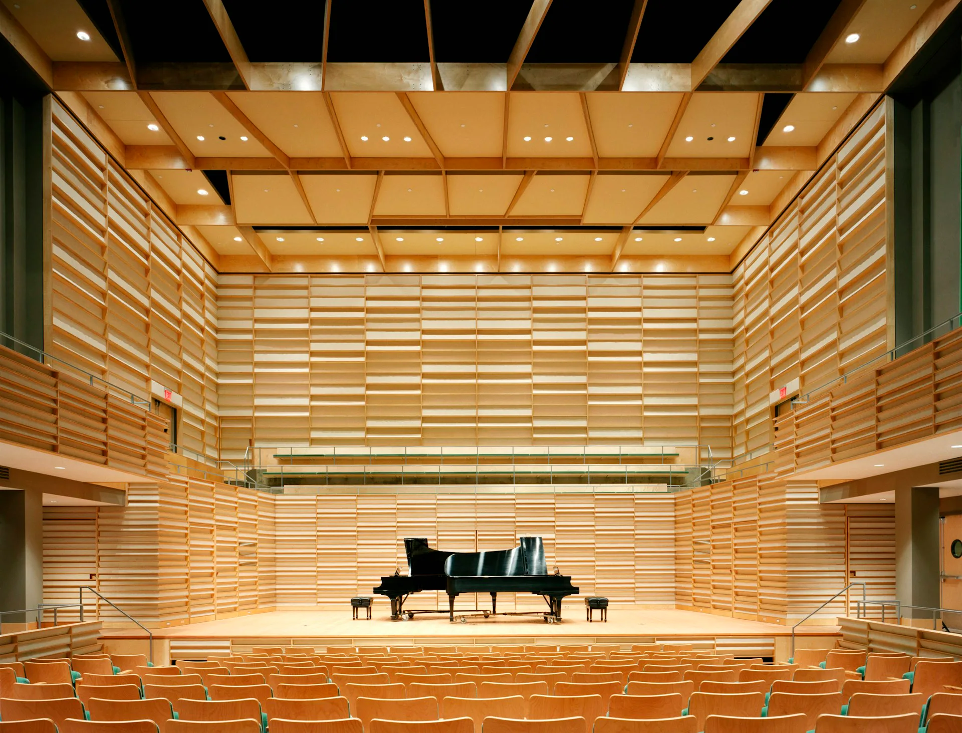 Modern concert hall interior with warm wood slat walls and ceiling, grand piano on stage, tiered seating, and integrated lighting by PKSB Architects.