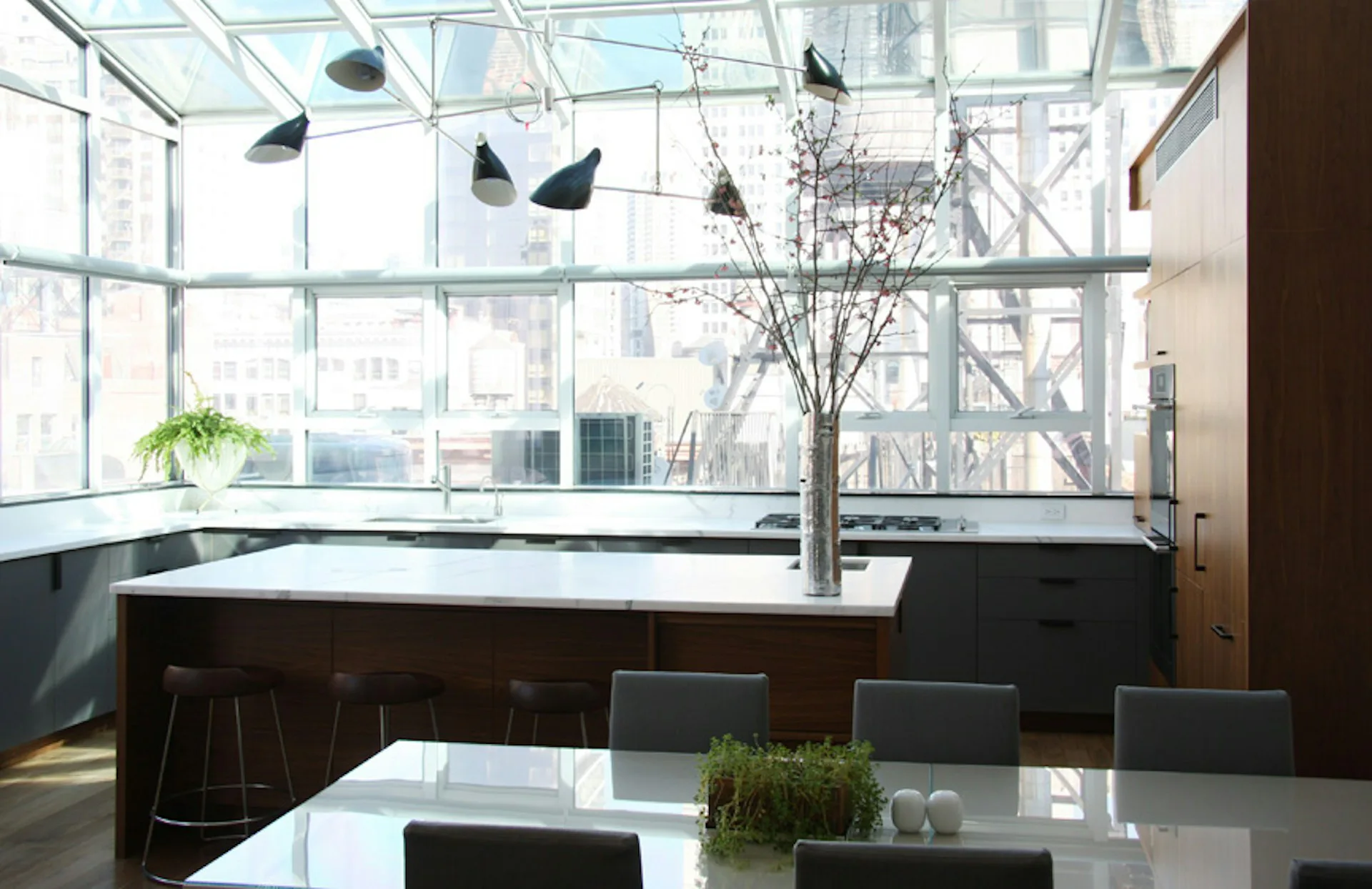 Modern loft kitchen with expansive gridded windows, dark cabinetry, white countertops, and hanging pendant lights overlooking a city skyline by Platt Dana Architects.