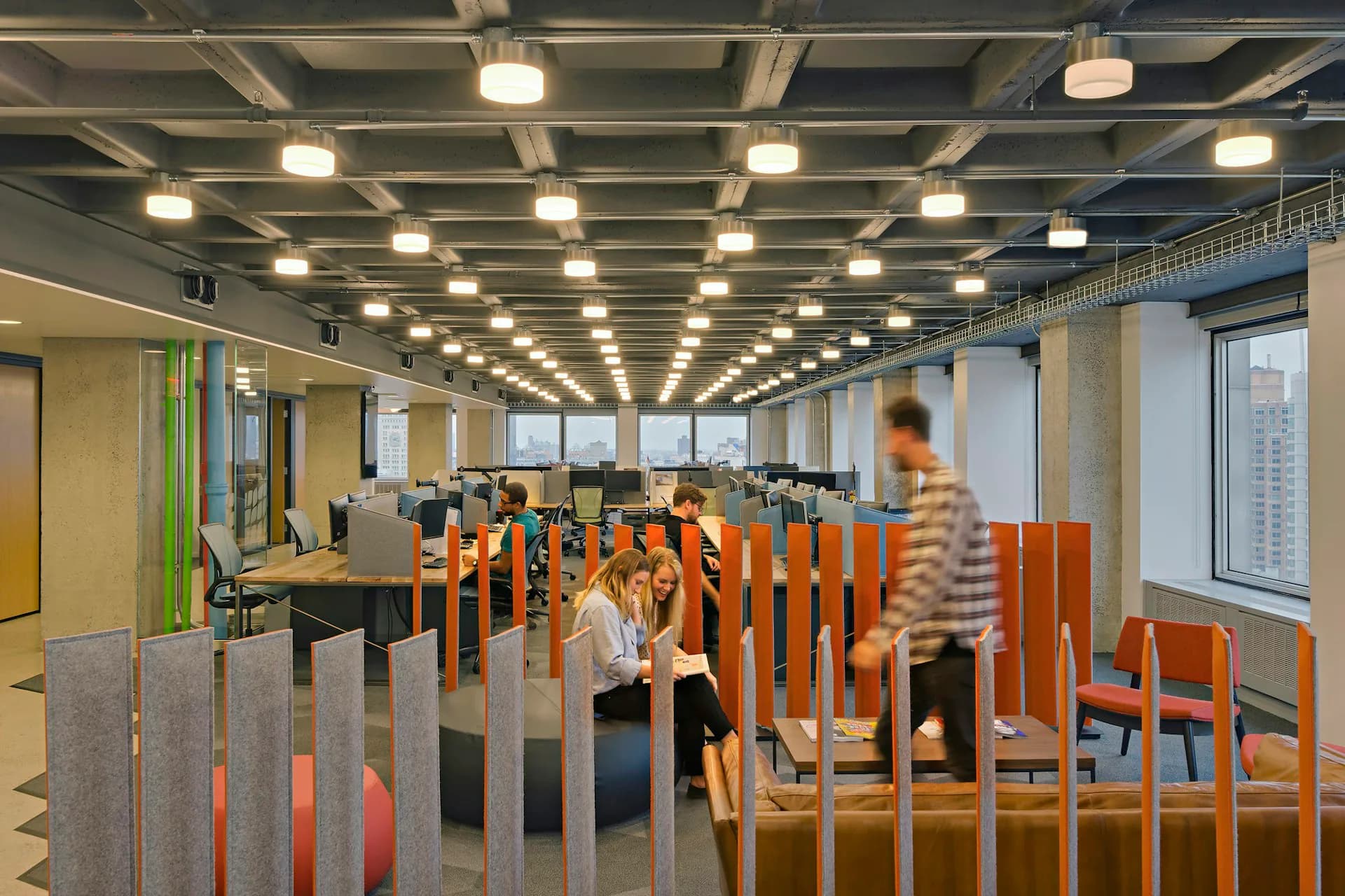 Modern open-plan office with orange wooden partition screens, bright ceiling lights, and views to working areas in the background by Pliskin Architecture PLLC.