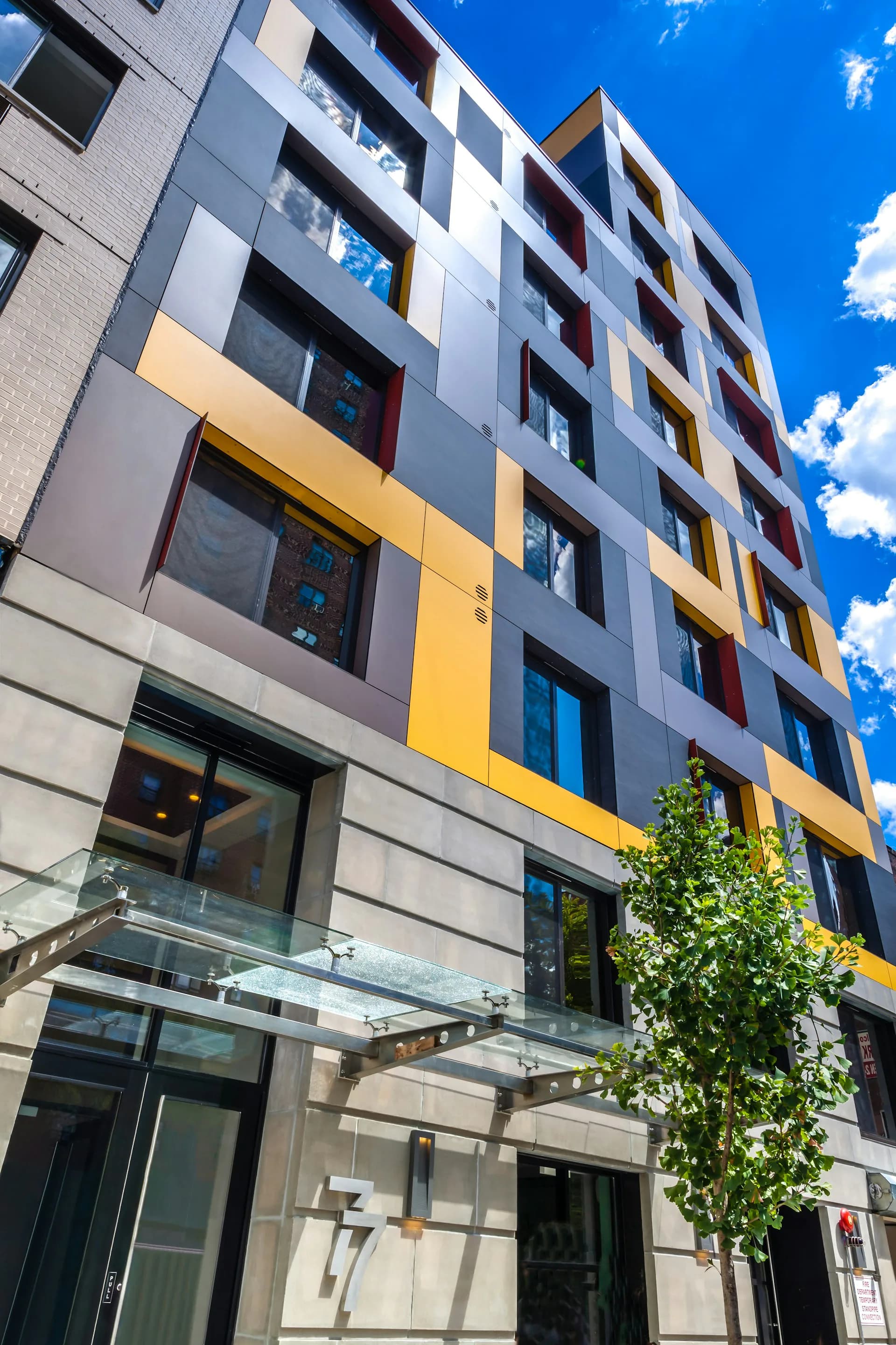 Modern residential building facade featuring white and gray cladding with yellow accent panels, glass balconies, and dark window frames against blue sky, by PM Architecture PC.