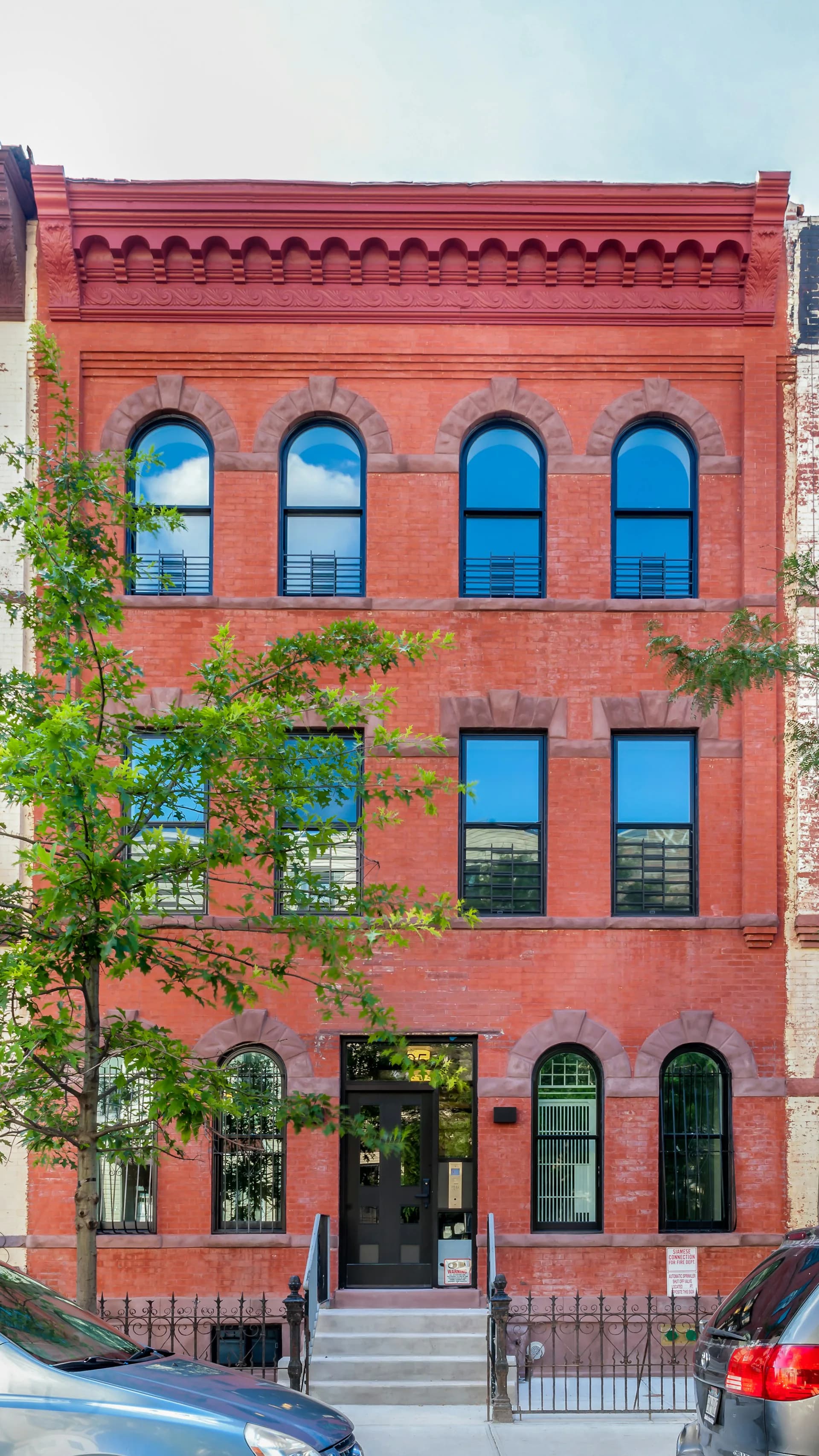 Red brick historic facade with blue-framed arched windows across three stories, decorative cornice, and climbing ivy by PM Architecture PC.