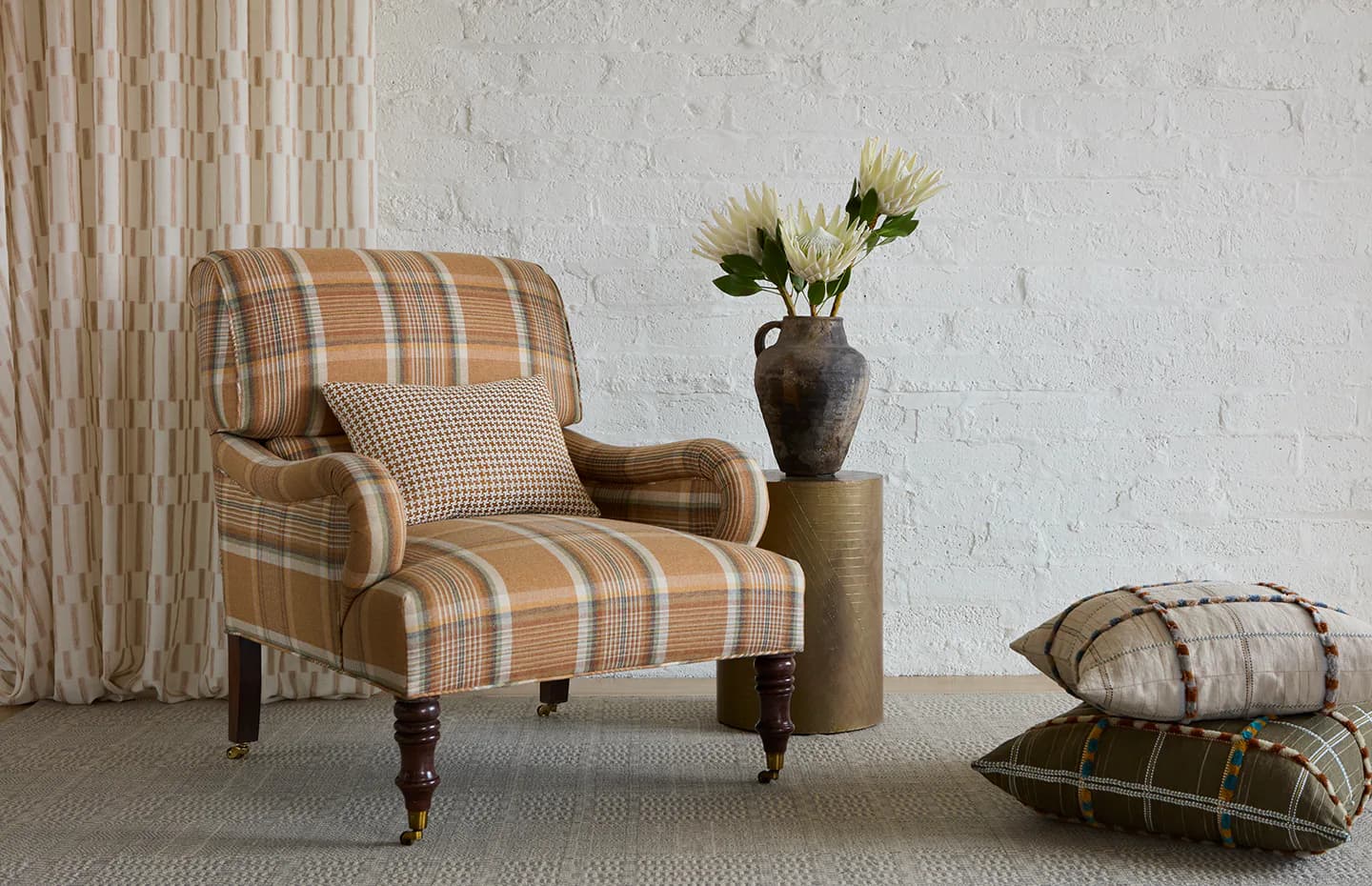 A stylized interior vignette featuring a plaid upholstered armchair, decorative pillows, flowers in a ceramic vase, and neutral textiles by Pollack.