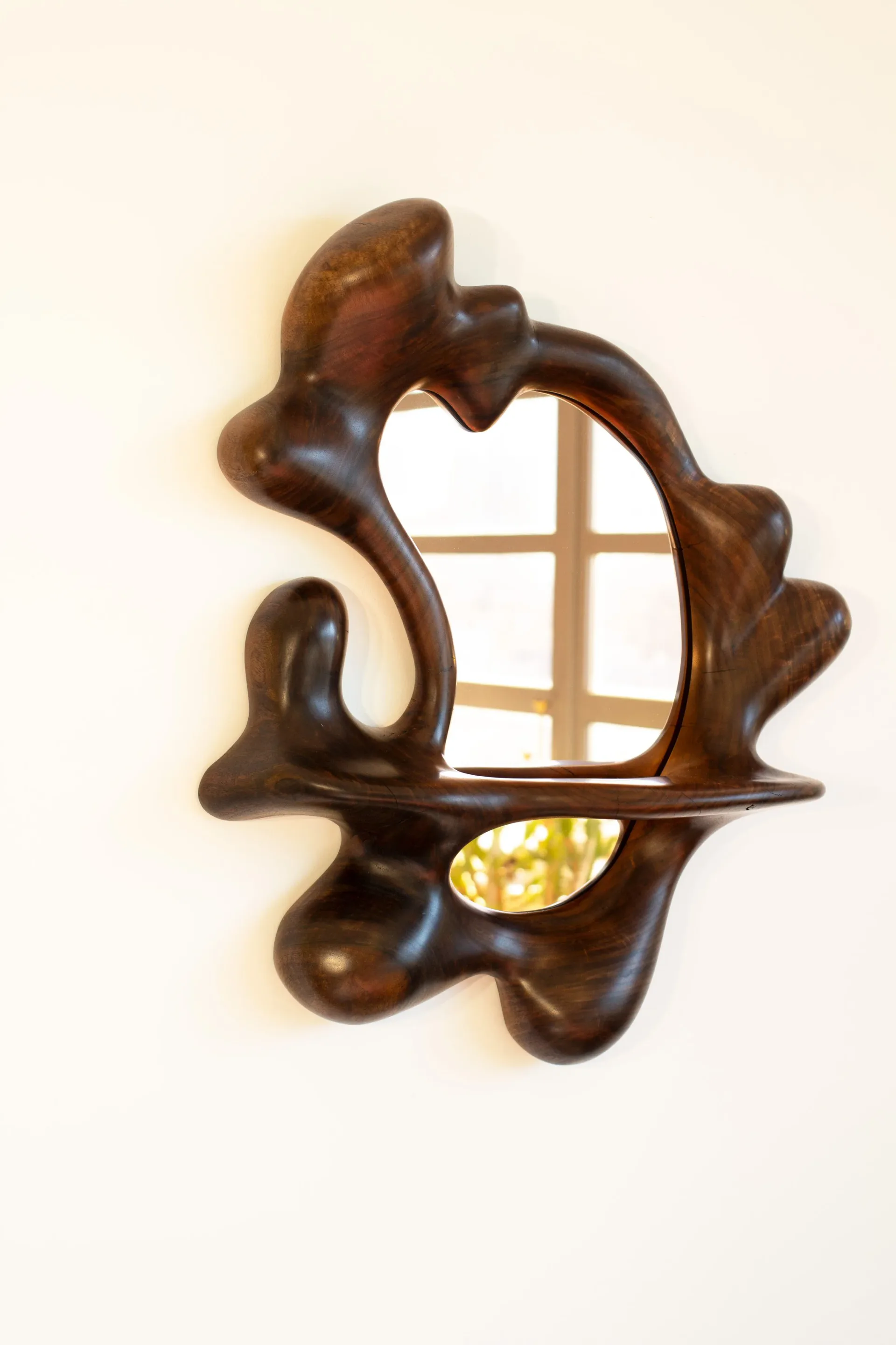 Dark wooden sculptural mirror with organic curved frame and grid-patterned glass center, mounted on white wall by Poritz & Studio.