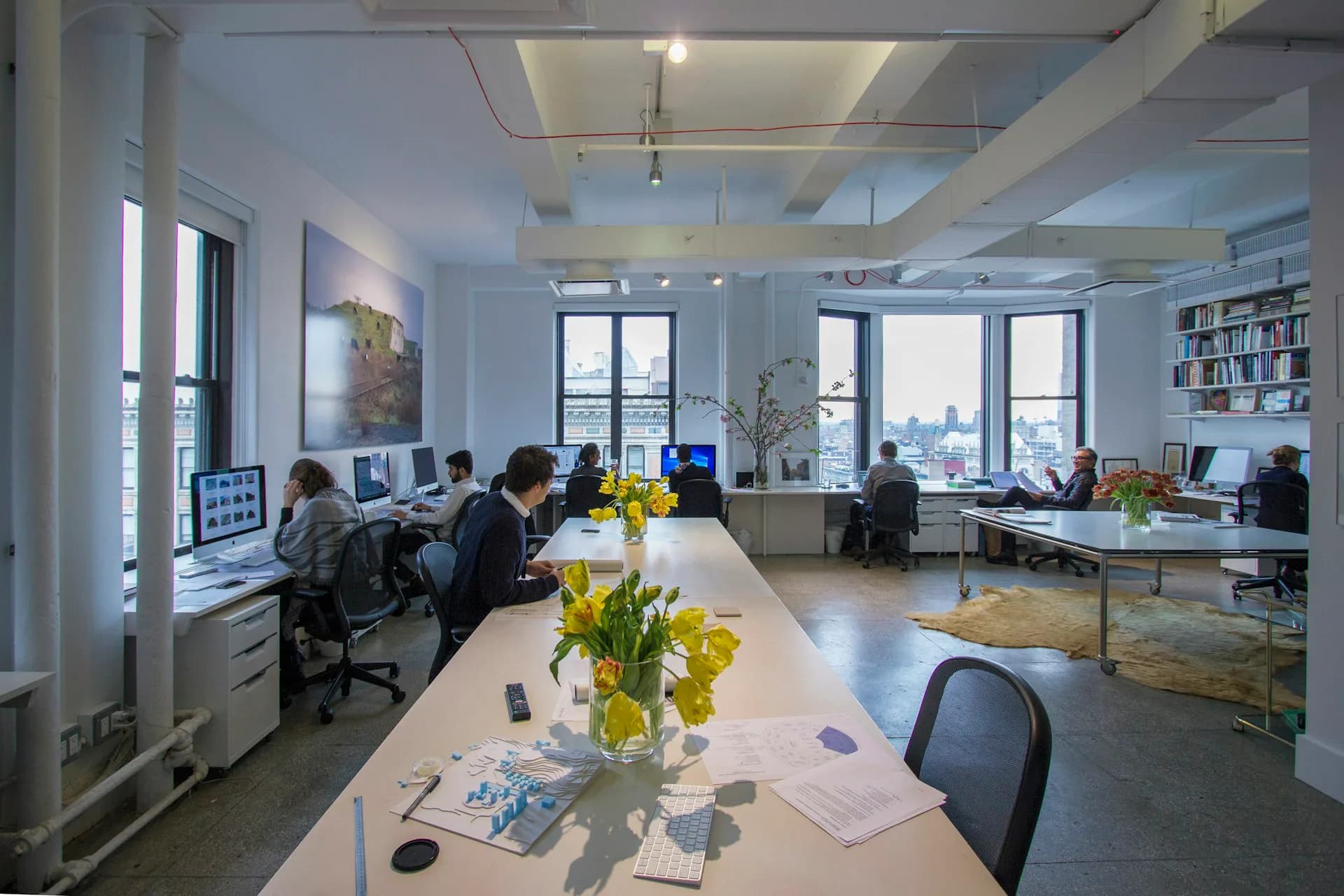 Bright modern office space with white walls, long communal table with yellow flowers, multiple workstations with monitors, large windows overlooking cityscape by Practice for Architecture and Urbanism.