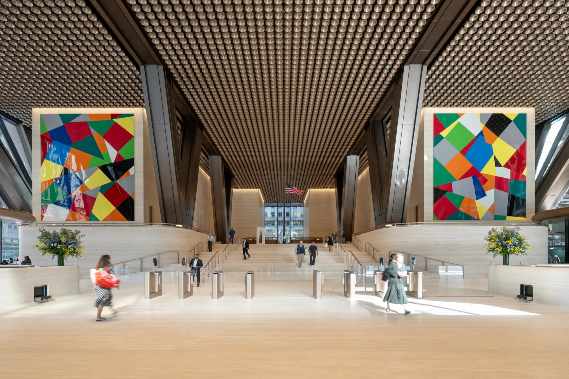 A spacious modern lobby with a geometric lattice ceiling, symmetrical colorful geometric artworks on side walls, light wood flooring, and minimalist furnishings by Practice for Architecture and Urbanism.