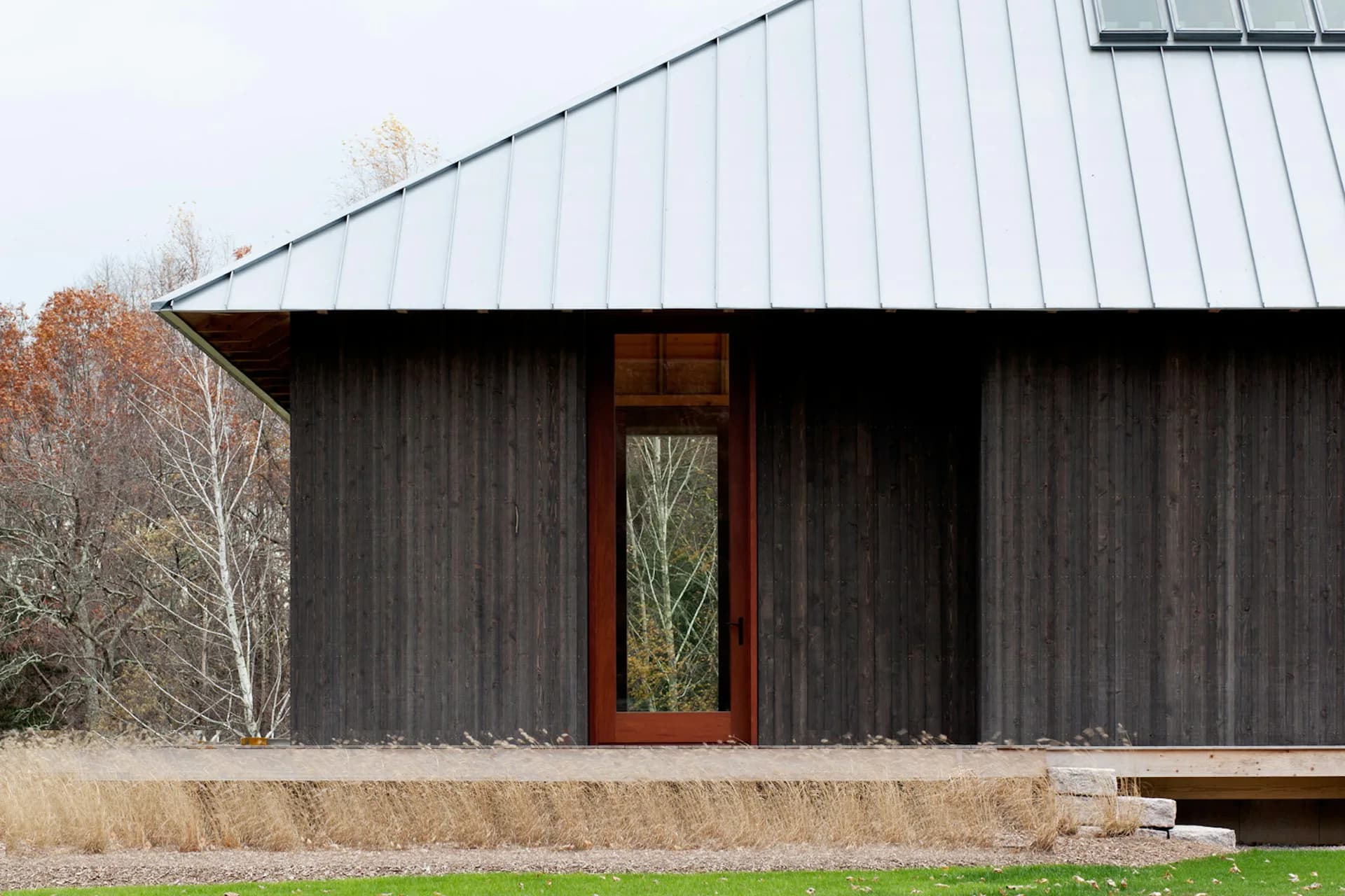 Dark corrugated metal residential building with warm wood-framed entry door and metal roof, set in a minimal landscape by P.R.O. - Peterson Rich Office.