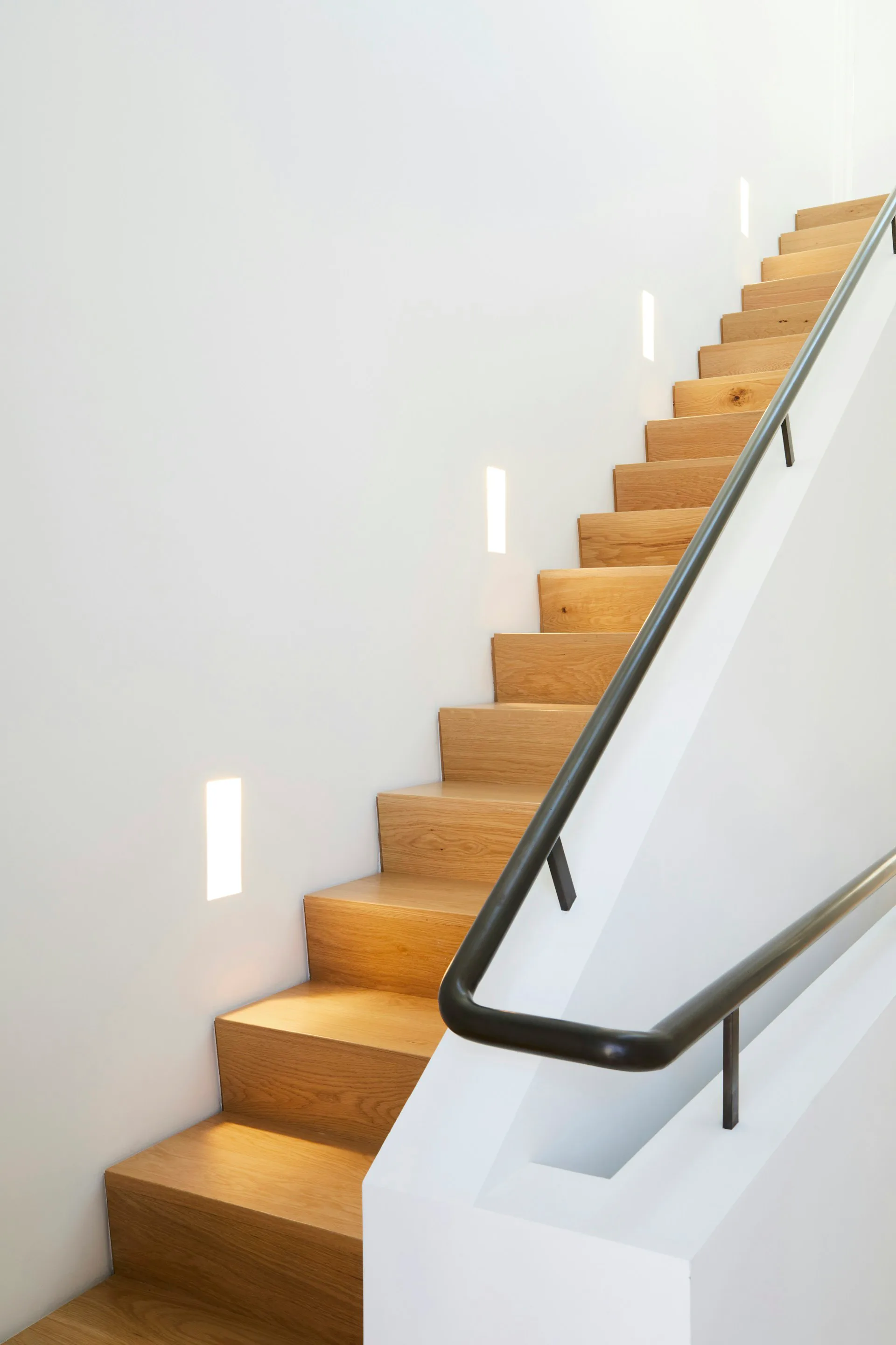Wooden staircase with black metal handrail ascending along white walls with integrated linear lighting by P.R.O. - Peterson Rich Office.