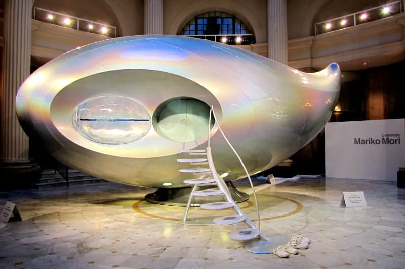 Futuristic sculptural pod installation with metallic iridescent finish and circular porthole window in gallery space by Prototype New York.