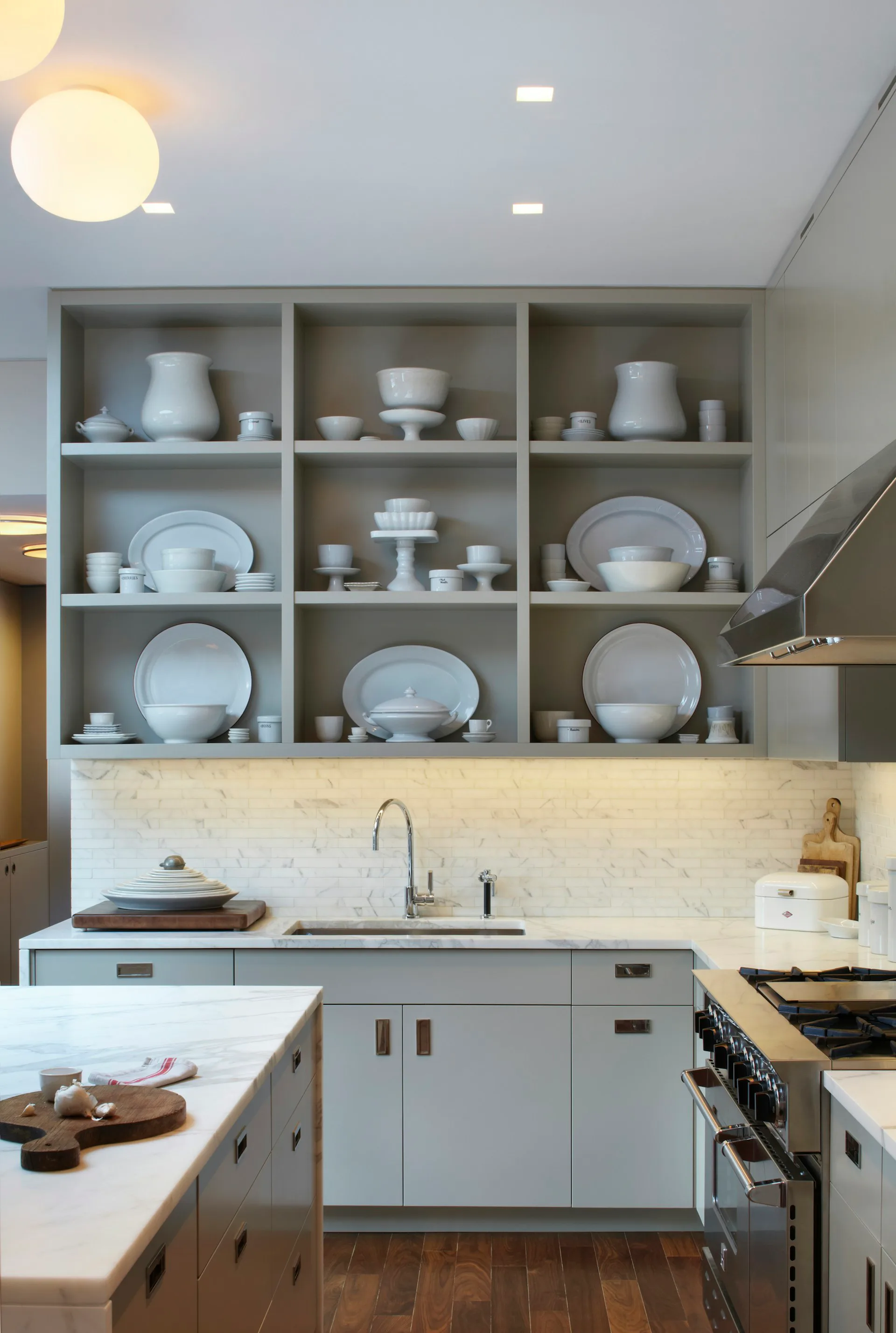 Modern kitchen with gray cabinetry, marble backsplash, and open shelving displaying white dishware and tableware by pulltab.
