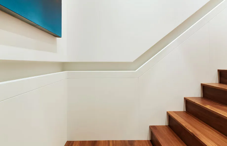 Modern staircase with wooden steps and white walls featuring integrated linear ceiling lighting and a teal accent panel, designed by pulltab.