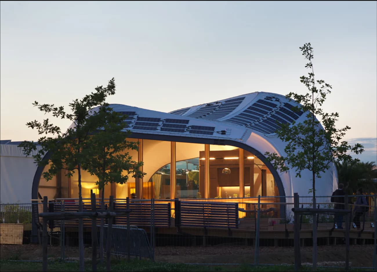 Modern curved pavilion with blue roof and expansive glazed walls, illuminated at dusk with surrounding landscape plantings by Pvilion.