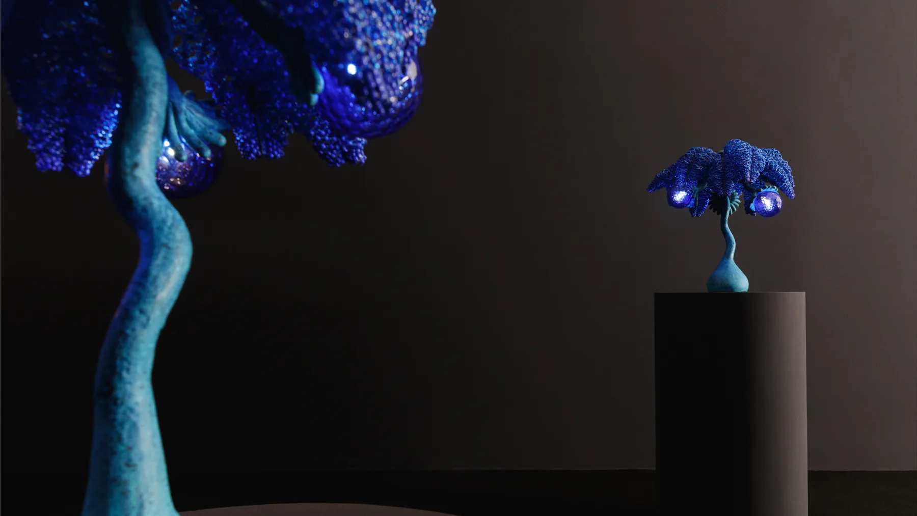 Two luminescent blue sculptural trees with glowing foliage against a dark background, one large on left and one smaller on pedestal on right, by R & Company.