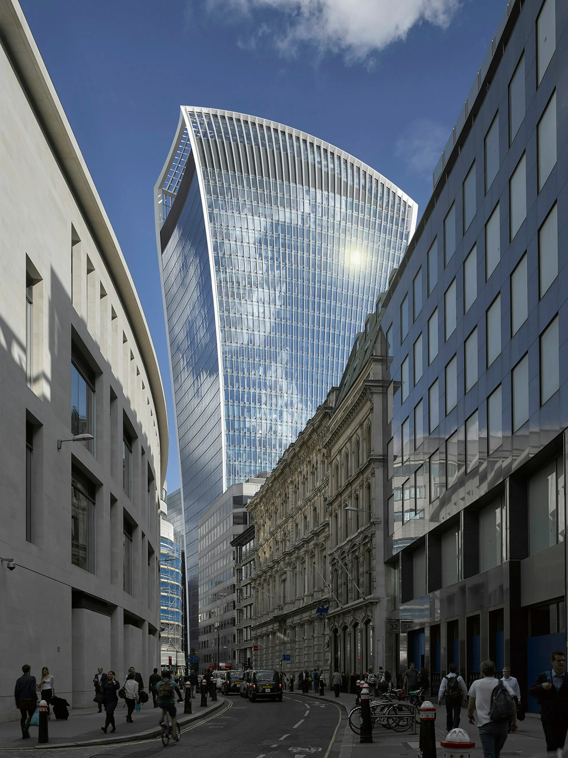 Modern curved glass skyscraper towers above surrounding historic stone buildings on a London street, featuring reflective facade and distinctive architectural design by Rafael Vinoly Architects.
