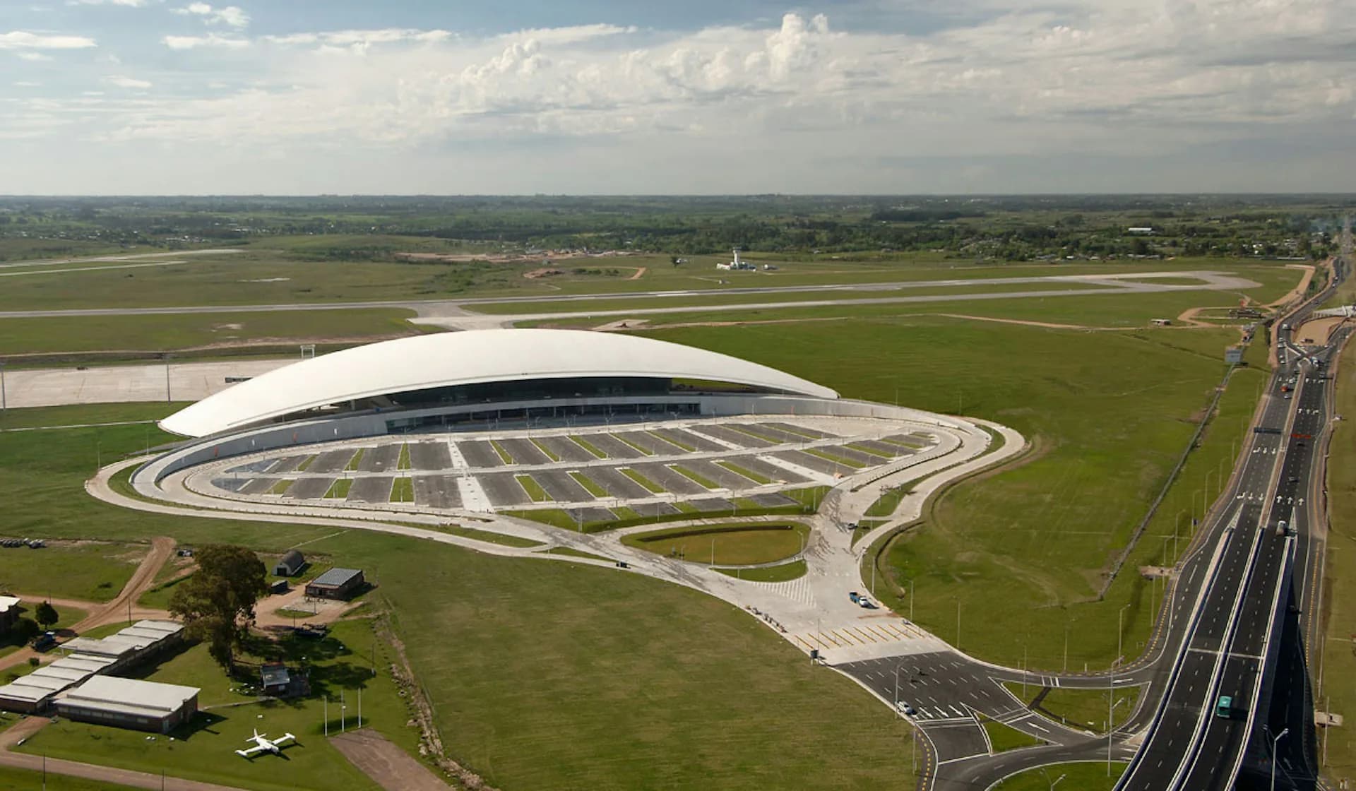 Aerial view of a modern oval-shaped stadium with white roof structure nestled in green agricultural landscape, by Rafael Vinoly Architects.